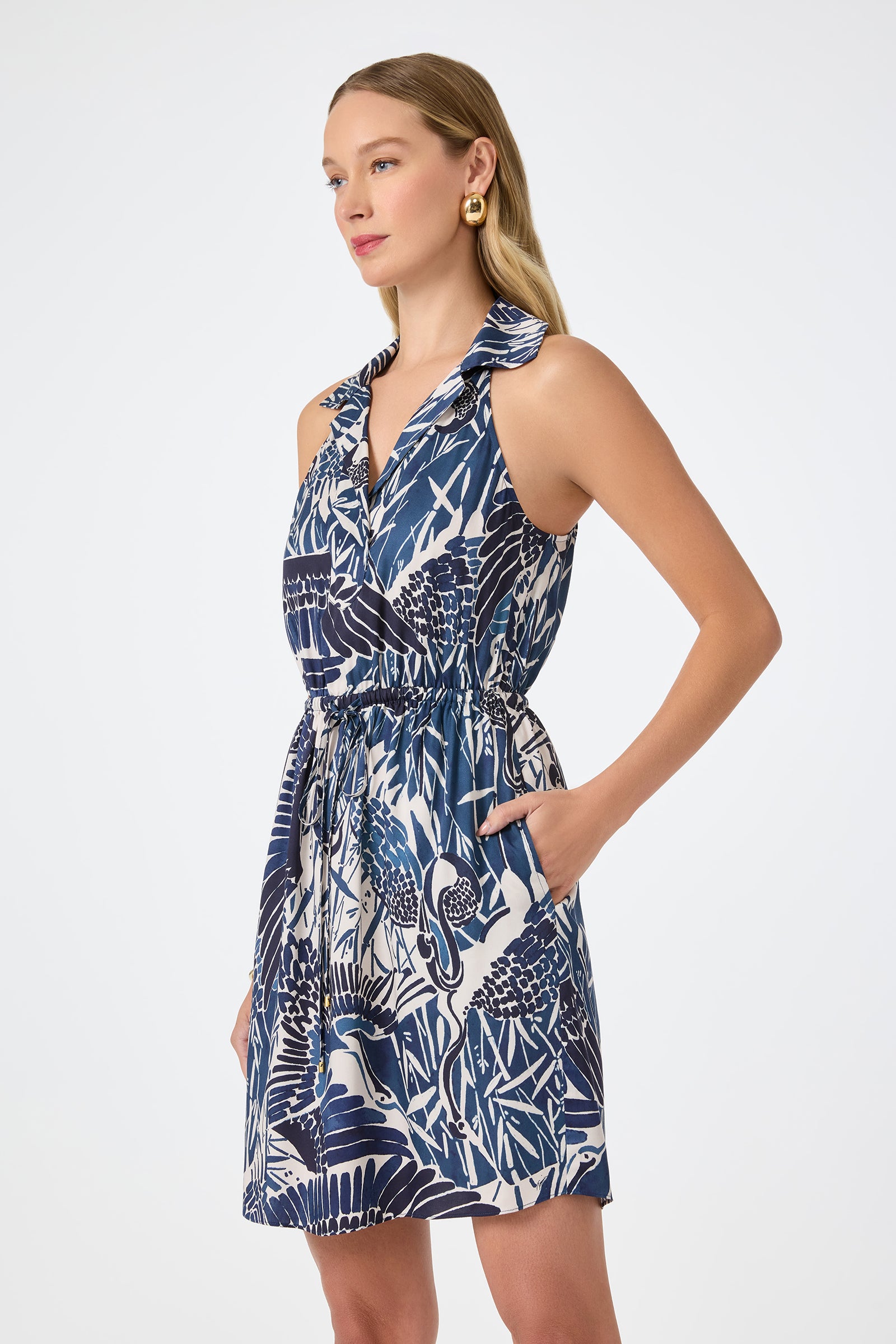 A woman models the Tsuru Caribbean 2 Mini Dress, a blue and white sleeveless dress with a ruffled collar and cinched waist, posing with one hand in her pocket against a plain light background.