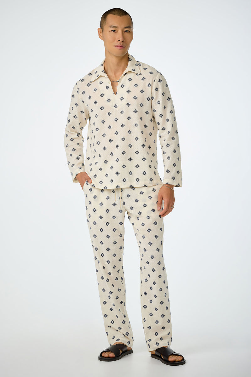 A person stands against a plain background wearing the Felipe Drawstring Pant in cream with a small blue geometric pattern, paired with black sandals. Their hands are relaxed by their sides.