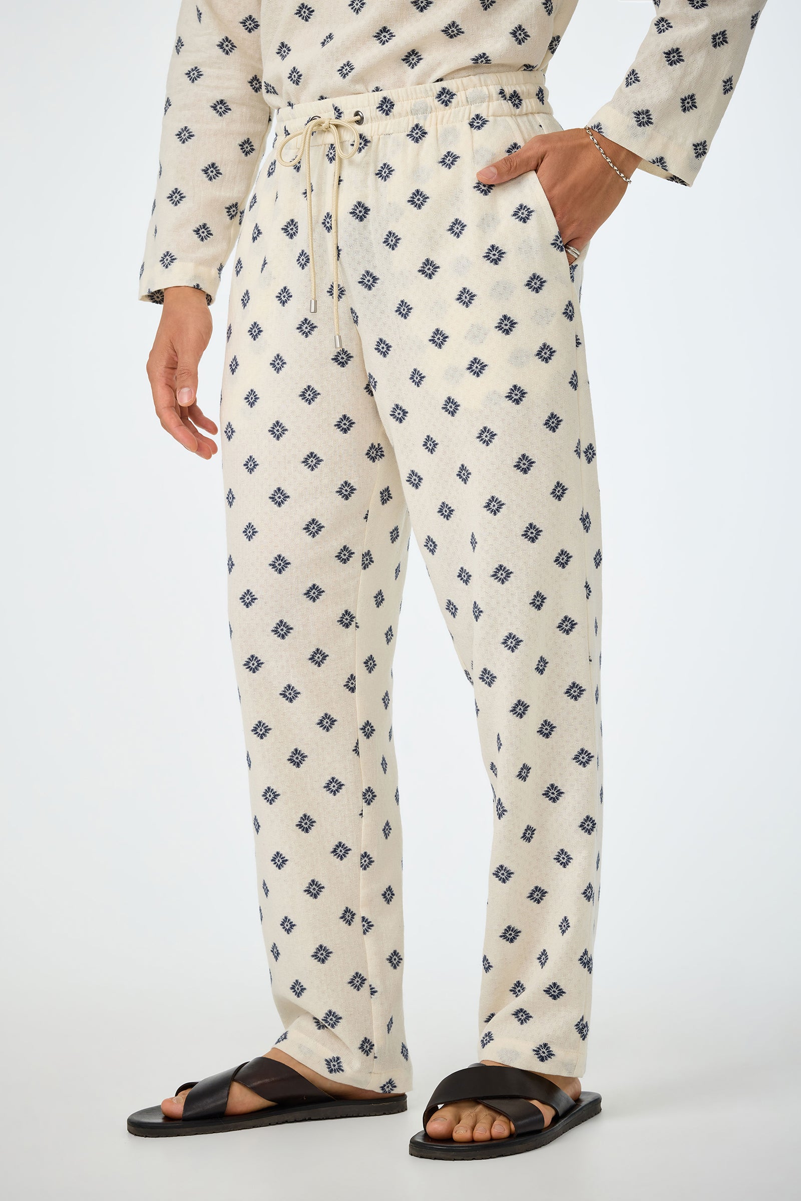 A person wears the Felipe Drawstring Pant in cream with a blue geometric diamond pattern, a matching top, black slide sandals, and a thin bracelet, posing with one hand in their pocket against a plain background.
