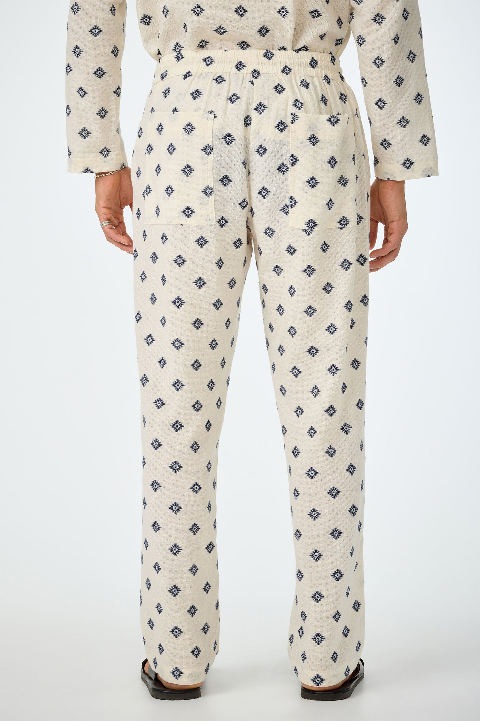 A person shown from the back wears Felipe Drawstring Pant in off-white with a blue geometric pattern, featuring an elastic waistband and relaxed fit, paired with matching long sleeves and sandals.