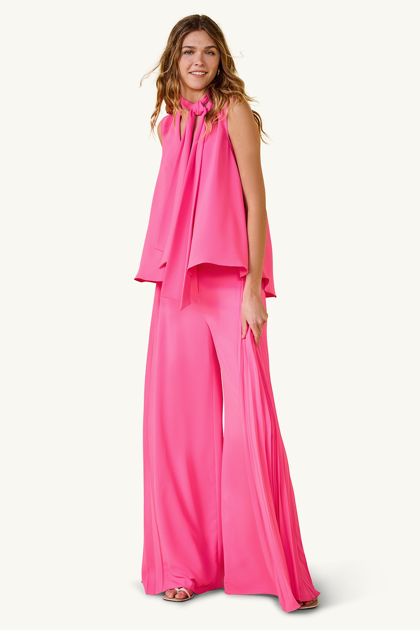 A woman wears the Frame Wide-Leg Pleated Pant in GS Georgette, styled with a vibrant hot pink sleeveless top featuring a long neck bow. She smiles with loose, wavy hair against a plain light background.