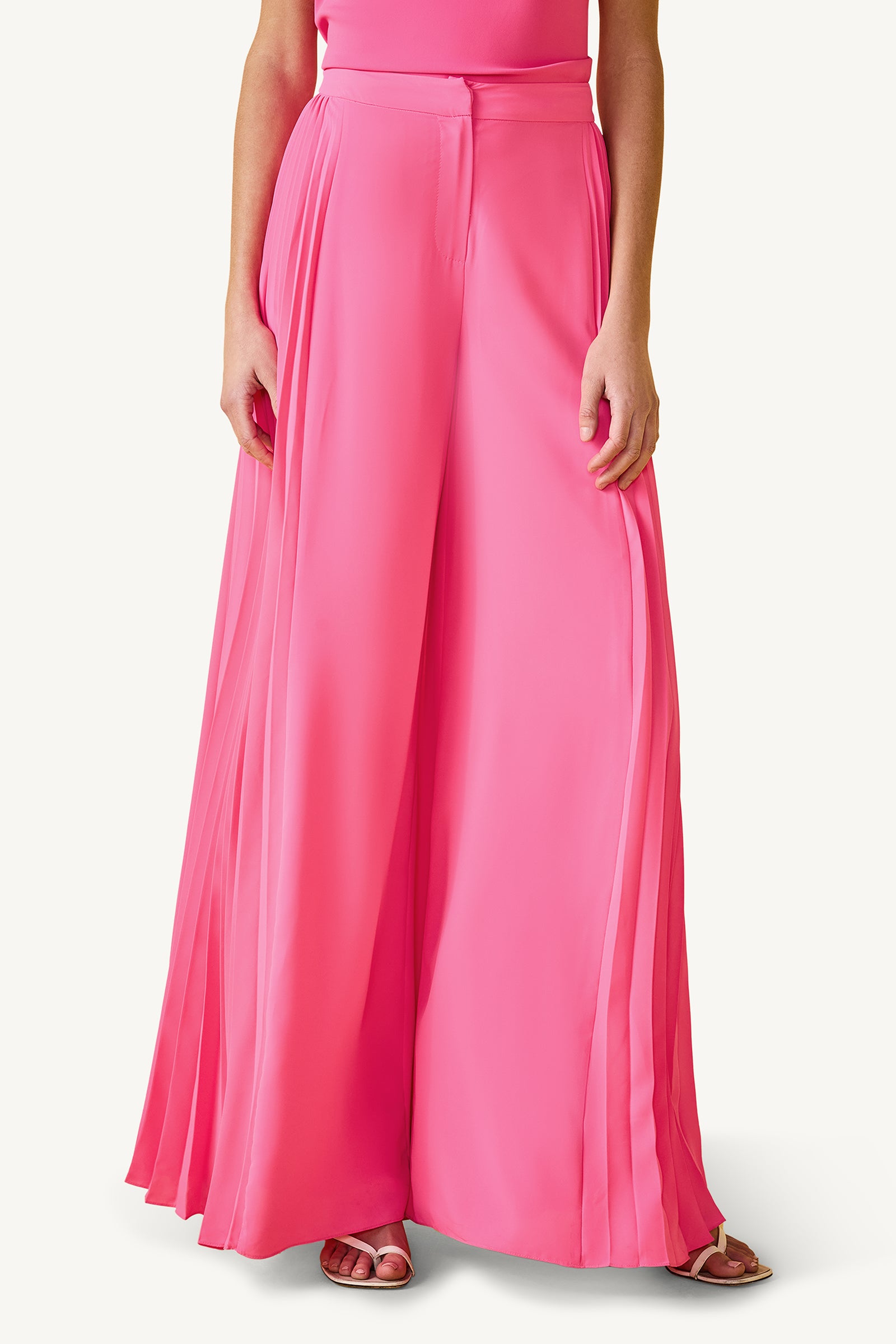Someone wears the Frame Wide-Leg Pleated Pant in Love All Pink GS Georgette—a high-waisted, flowy, pleated style with wide legs—standing hands at sides and paired with open-toed shoes. Upper body is partially visible.