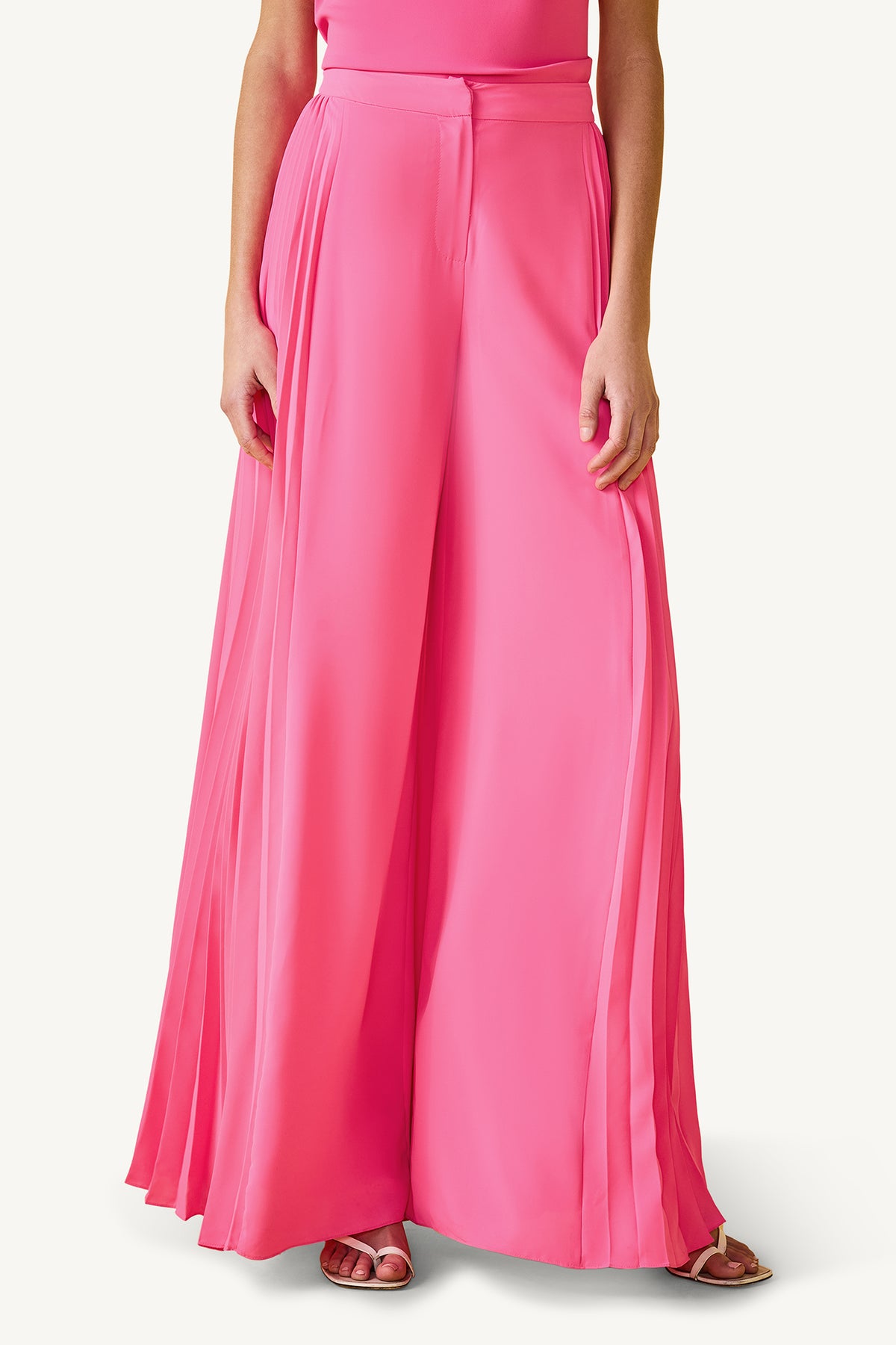 Someone wears the Frame Wide-Leg Pleated Pant in Love All Pink GS Georgette—a high-waisted, flowy, pleated style with wide legs—standing hands at sides and paired with open-toed shoes. Upper body is partially visible.