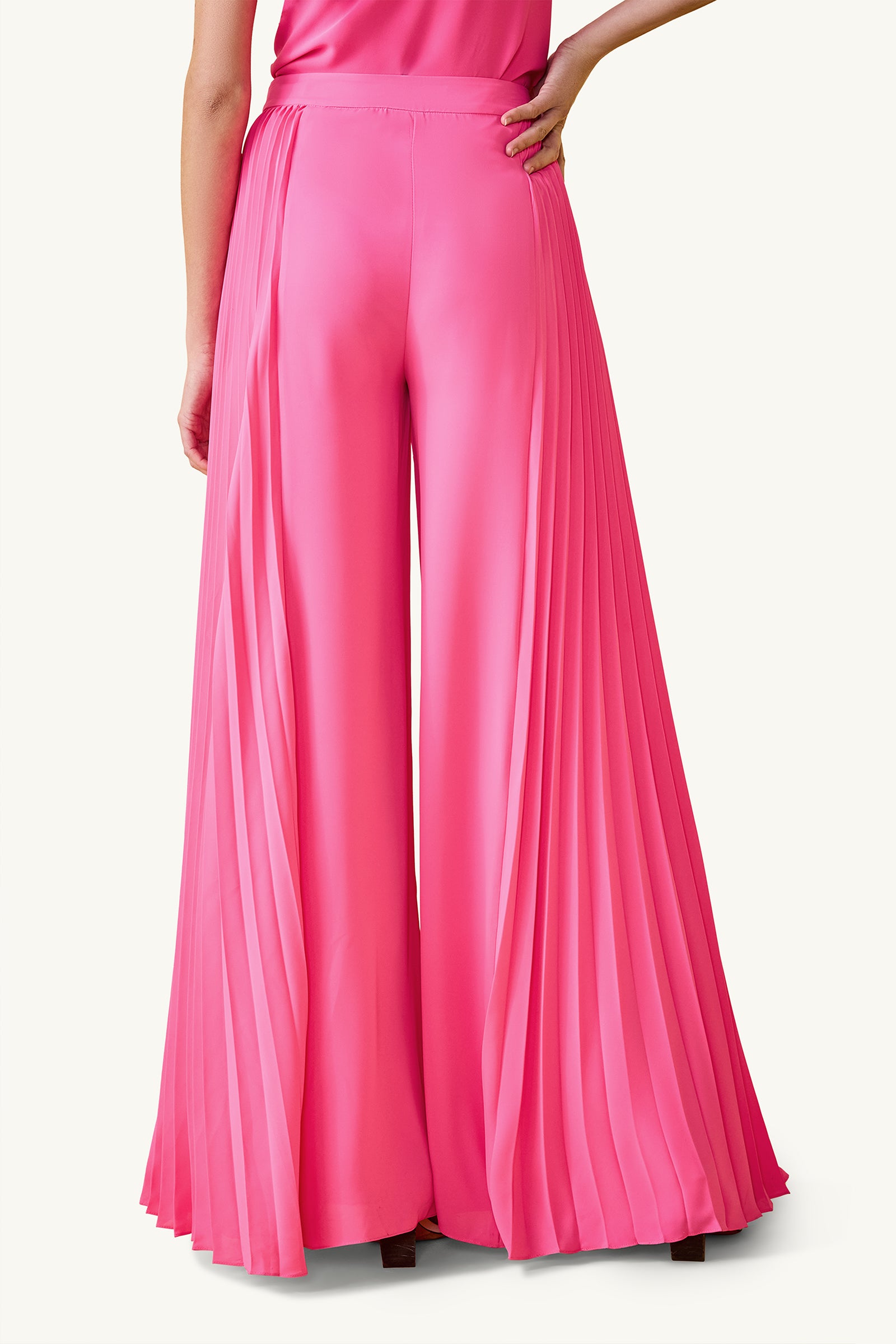 A person is seen from the back wearing Frame Wide-Leg Pleated Pant - GS Georgette in bright pink. The shiny, high-waisted pants feature pleats and wide legs that flow gracefully to the floor.