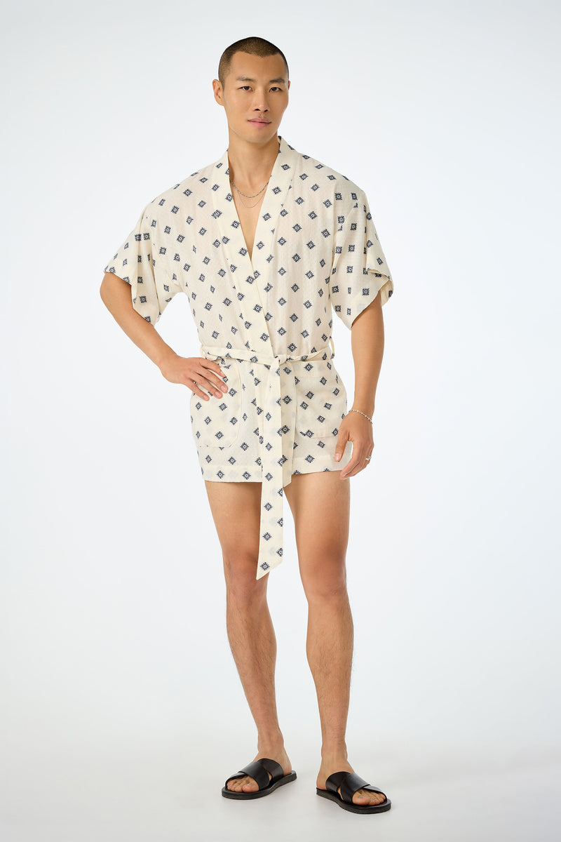 A man stands against a plain background wearing the Happi Coat—a short, cream-colored romper with a dark geometric pattern, short sleeves, a tie belt—paired with black sandals and gold jewelry.