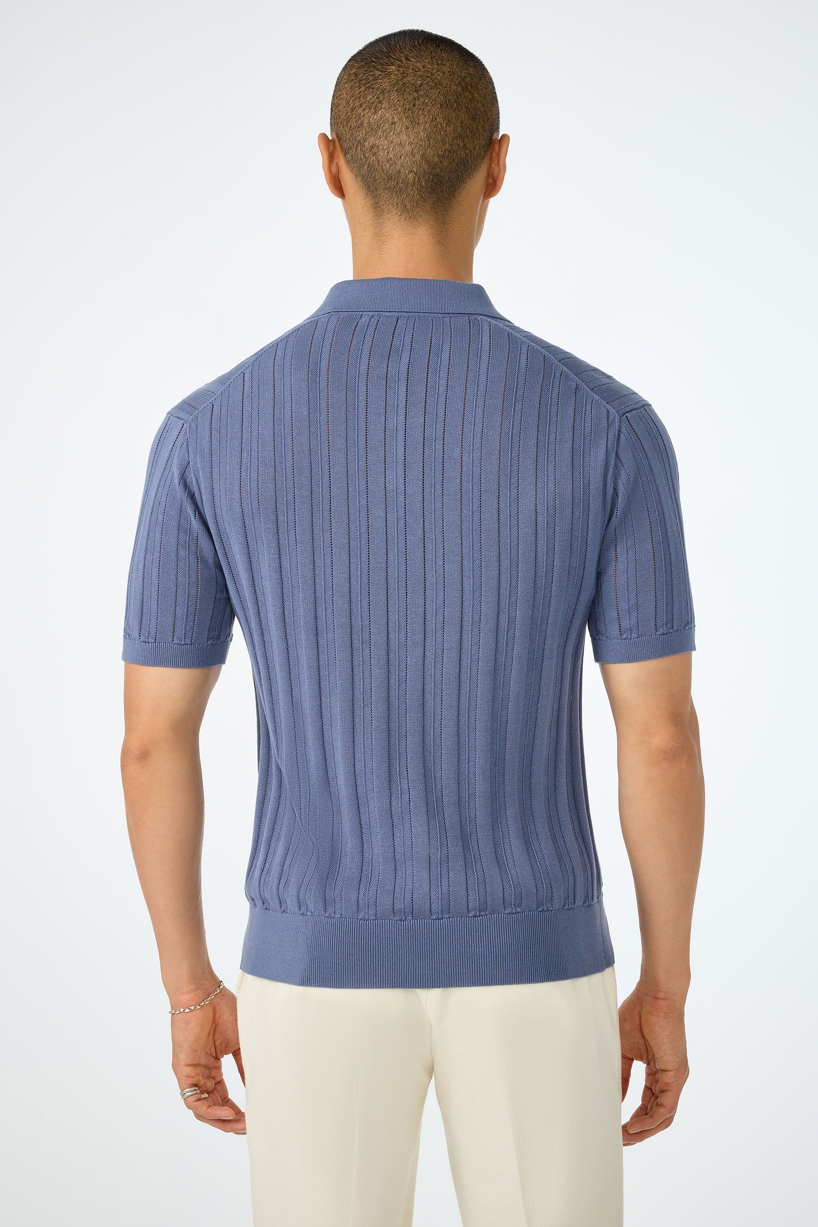 A person faces away, wearing the Kensington Short-Sleeve Polo in ribbed light blue knit and off-white pants, set against a plain light background.