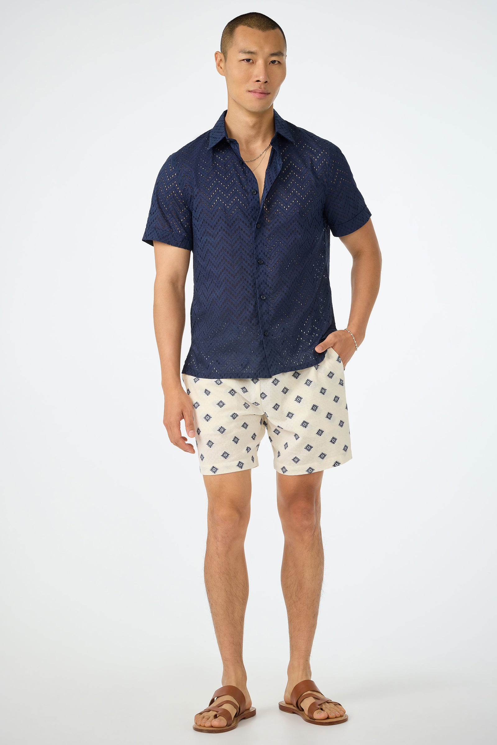 A man stands against a white background, wearing the Shoreline Short - Foulard Gauze, a navy chevron-patterned short-sleeve shirt, and brown sandals. One hand is in his pocket as he looks directly at the camera.