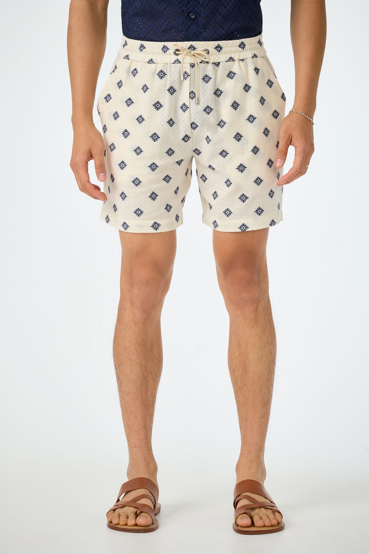 A person stands against a plain white background wearing the Shoreline Short - Foulard Gauze in ecru with a navy tucked-in shirt, brown sandals, and a bracelet.