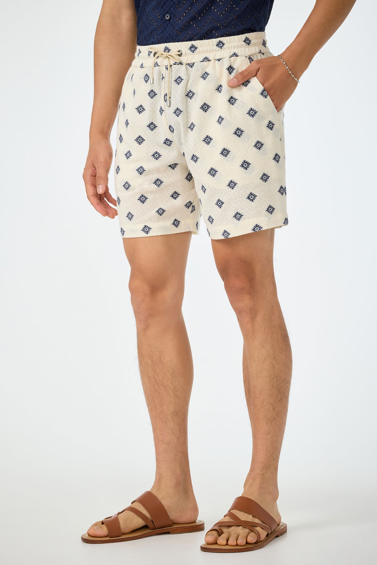 A person stands against a plain background wearing the Shoreline Short - Foulard Gauze, featuring a cream base with blue geometric pattern, paired with a navy shirt, brown sandals, and a silver bracelet, one hand in their pocket.