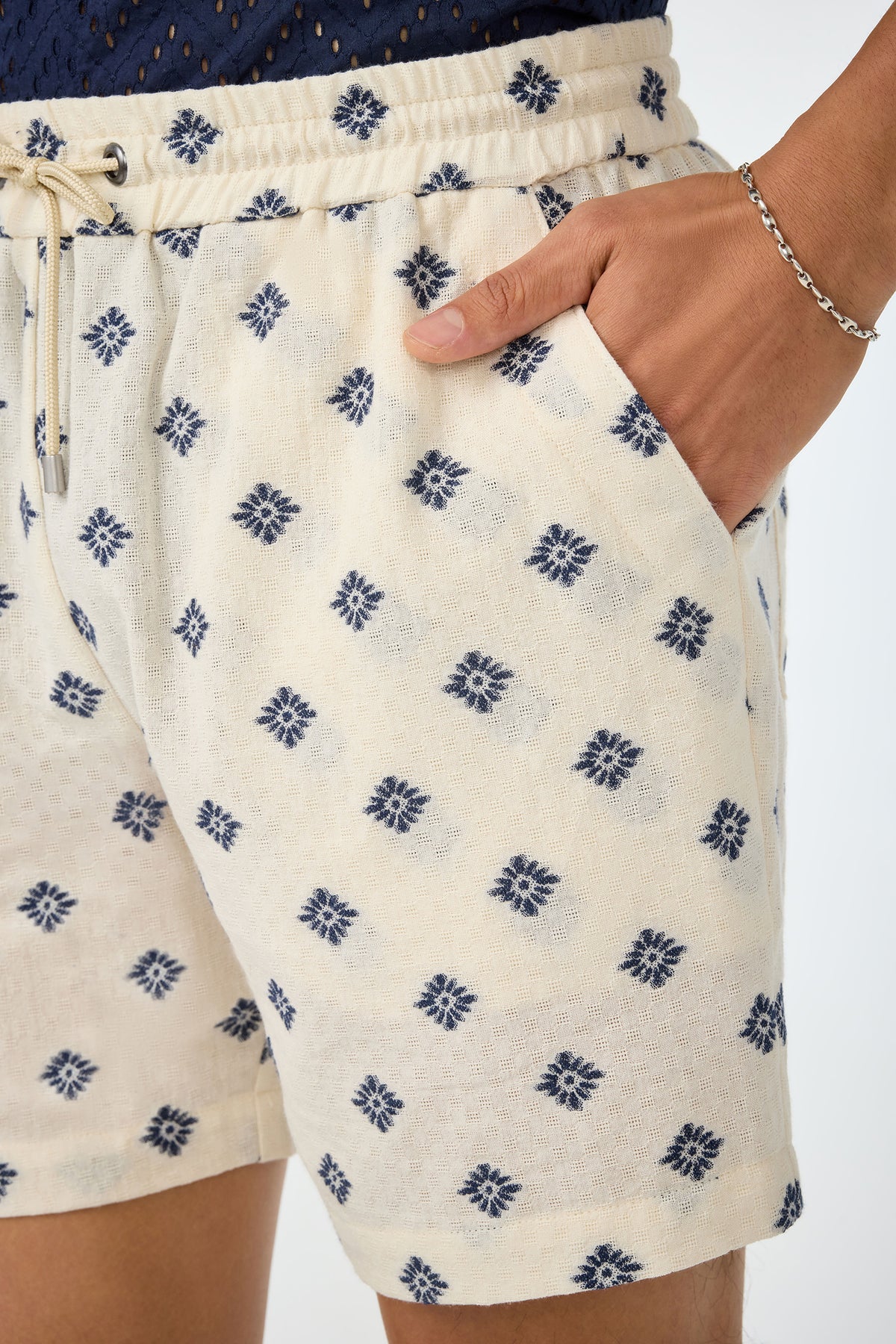 A person wears the Shoreline Short - Foulard Gauze, featuring a cream fabric with a blue geometric print, drawstring waist, and one hand in the pocket. A thin silver bracelet adorns their wrist.