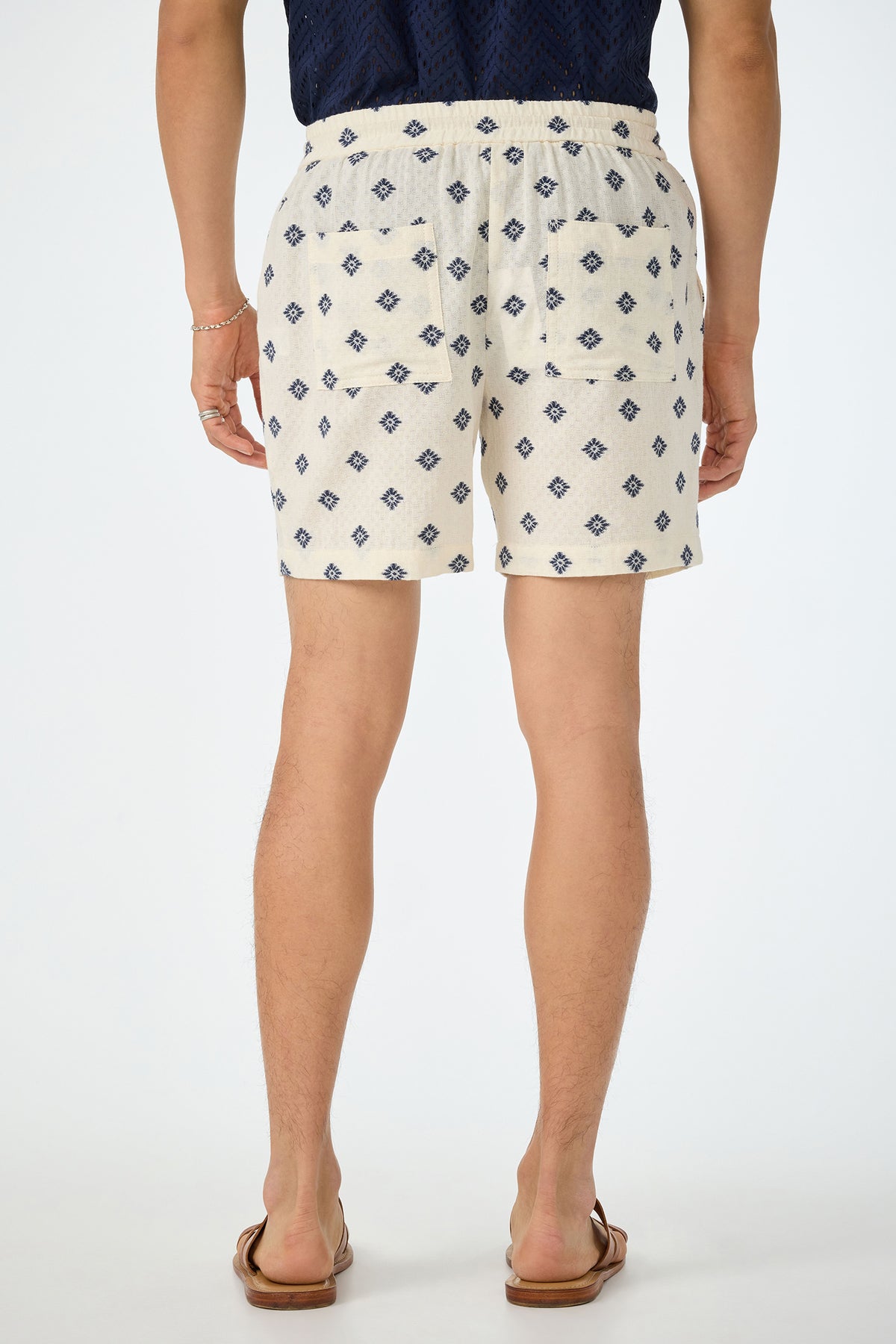 A person stands facing away, wearing the Shoreline Short - Foulard Gauze, featuring a cream base with a blue diamond pattern. They pair it with a navy shirt, brown sandals, and a bracelet. The background is plain white.