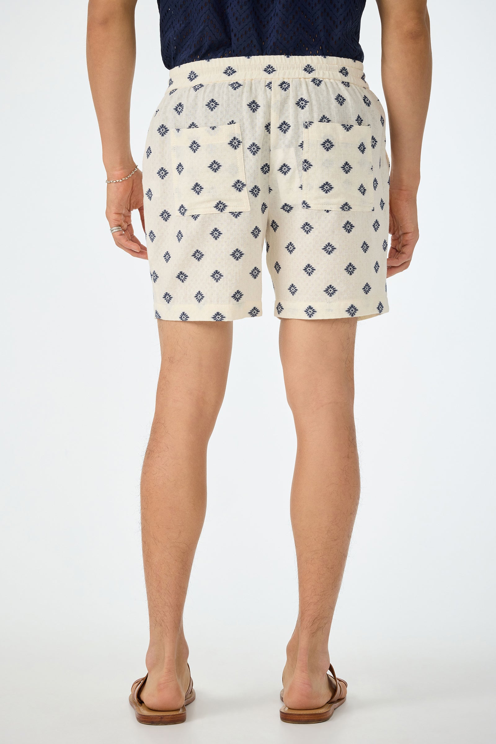 A person stands facing away, wearing the Shoreline Short - Foulard Gauze, featuring a cream base with a blue diamond pattern. They pair it with a navy shirt, brown sandals, and a bracelet. The background is plain white.