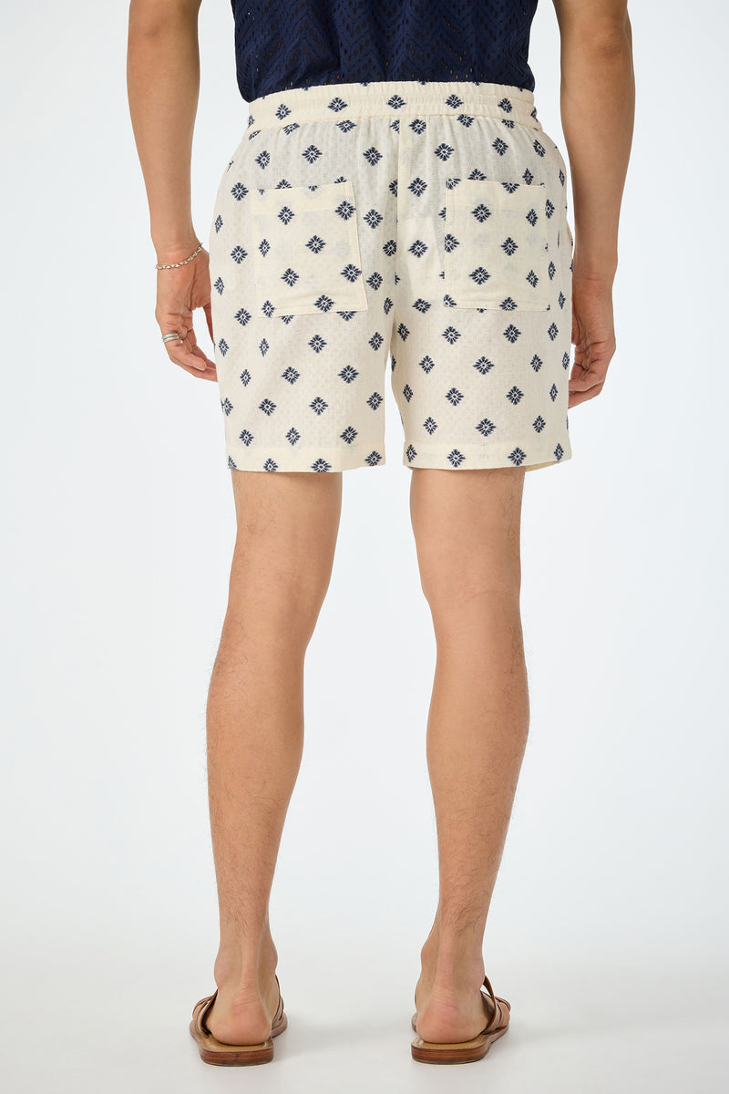 A person stands facing away, wearing the Shoreline Short - Foulard Gauze, featuring a cream base with a blue diamond pattern. They pair it with a navy shirt, brown sandals, and a bracelet. The background is plain white.
