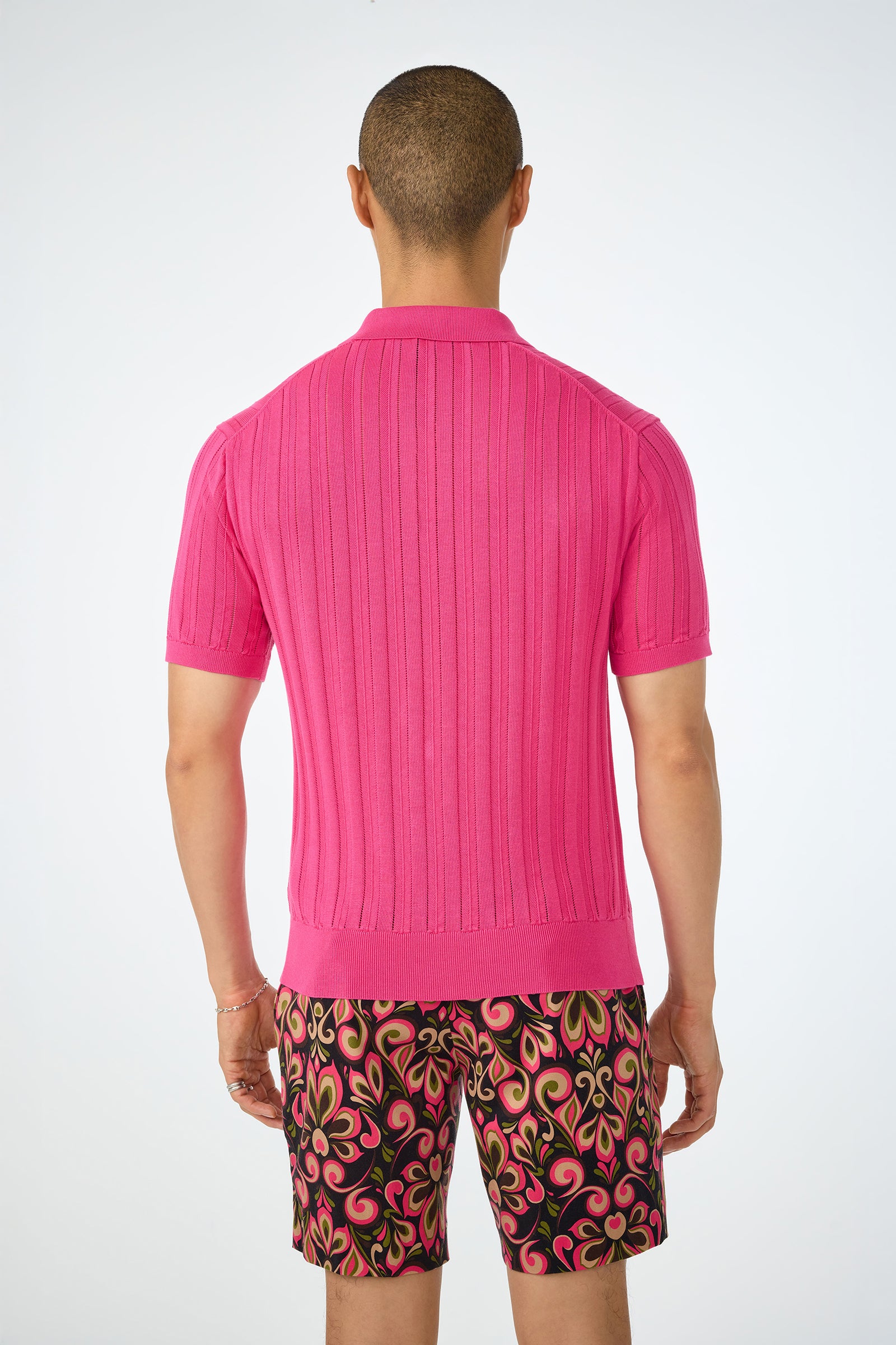 A person faces away, wearing the Kensington Short-Sleeve Polo in bright pink and patterned shorts with pink, yellow, and black designs, set against a plain light background.
