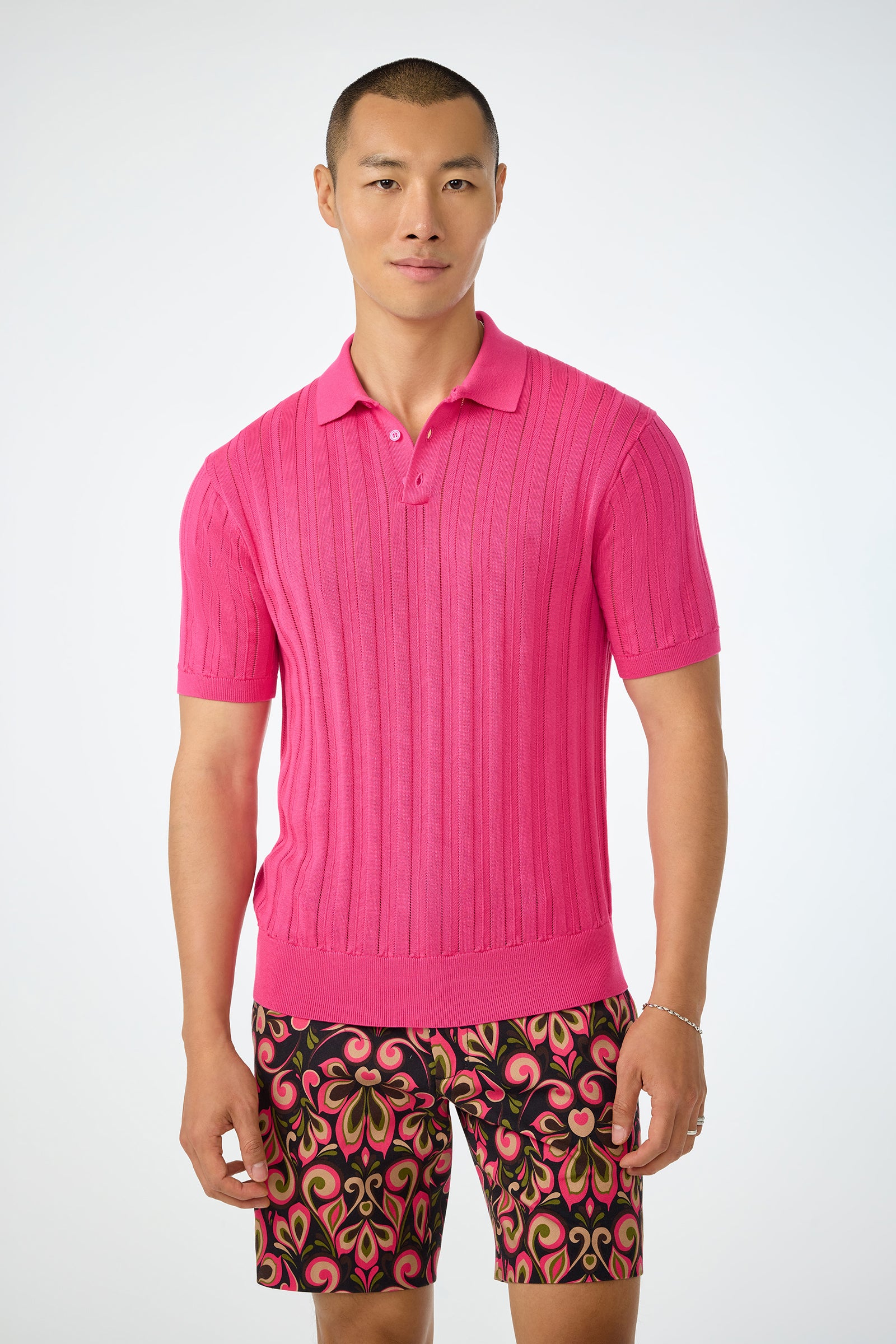 A man with short hair and a neutral expression stands against a plain background, wearing the Kensington Short-Sleeve Polo in Forrest Rose and patterned shorts featuring a colorful, swirling design.