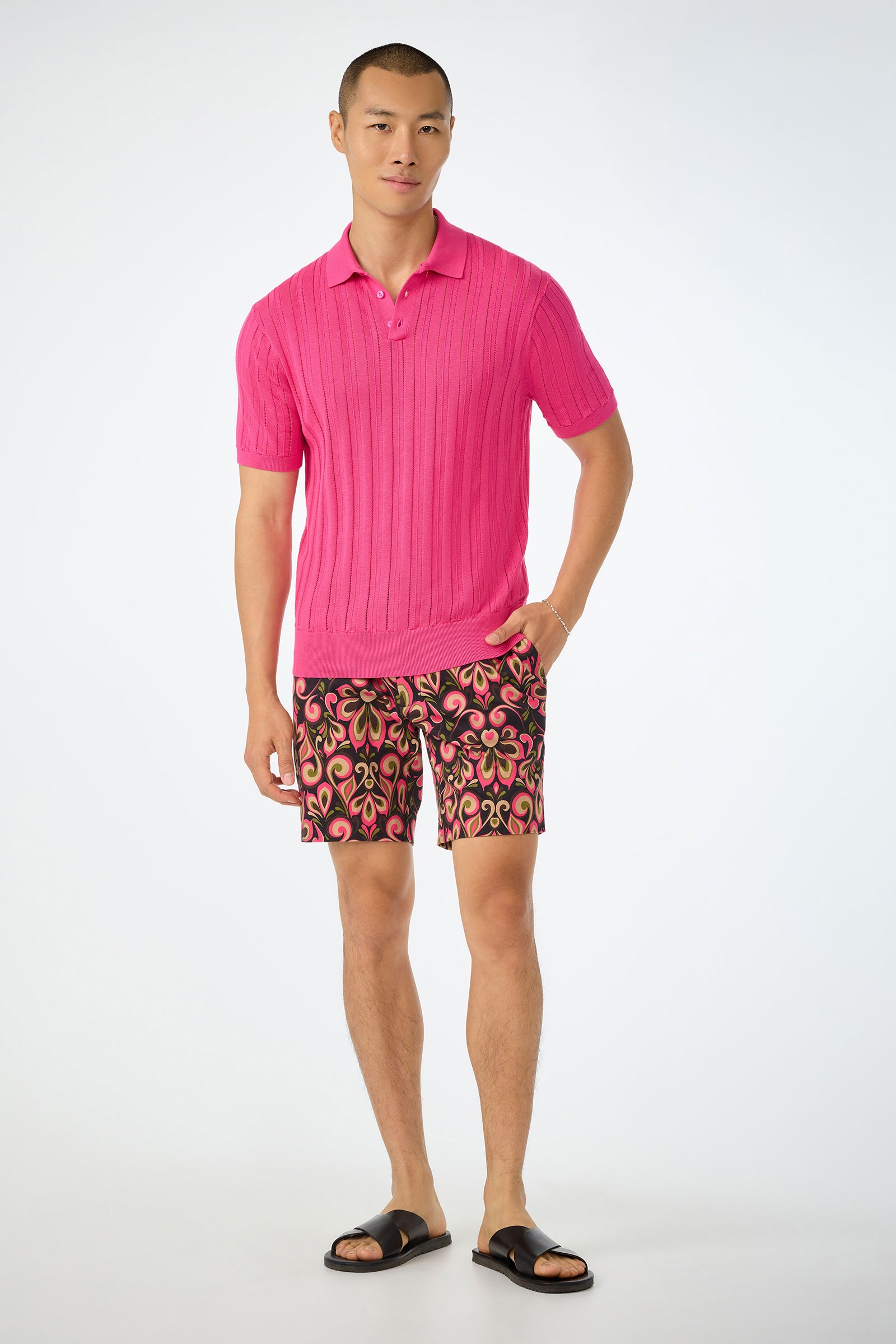 A man stands against a light background wearing the Kensington Short-Sleeve Polo in bright pink, colorful swirl-pattern shorts, black sandals, and has a relaxed expression with one hand in his pocket.