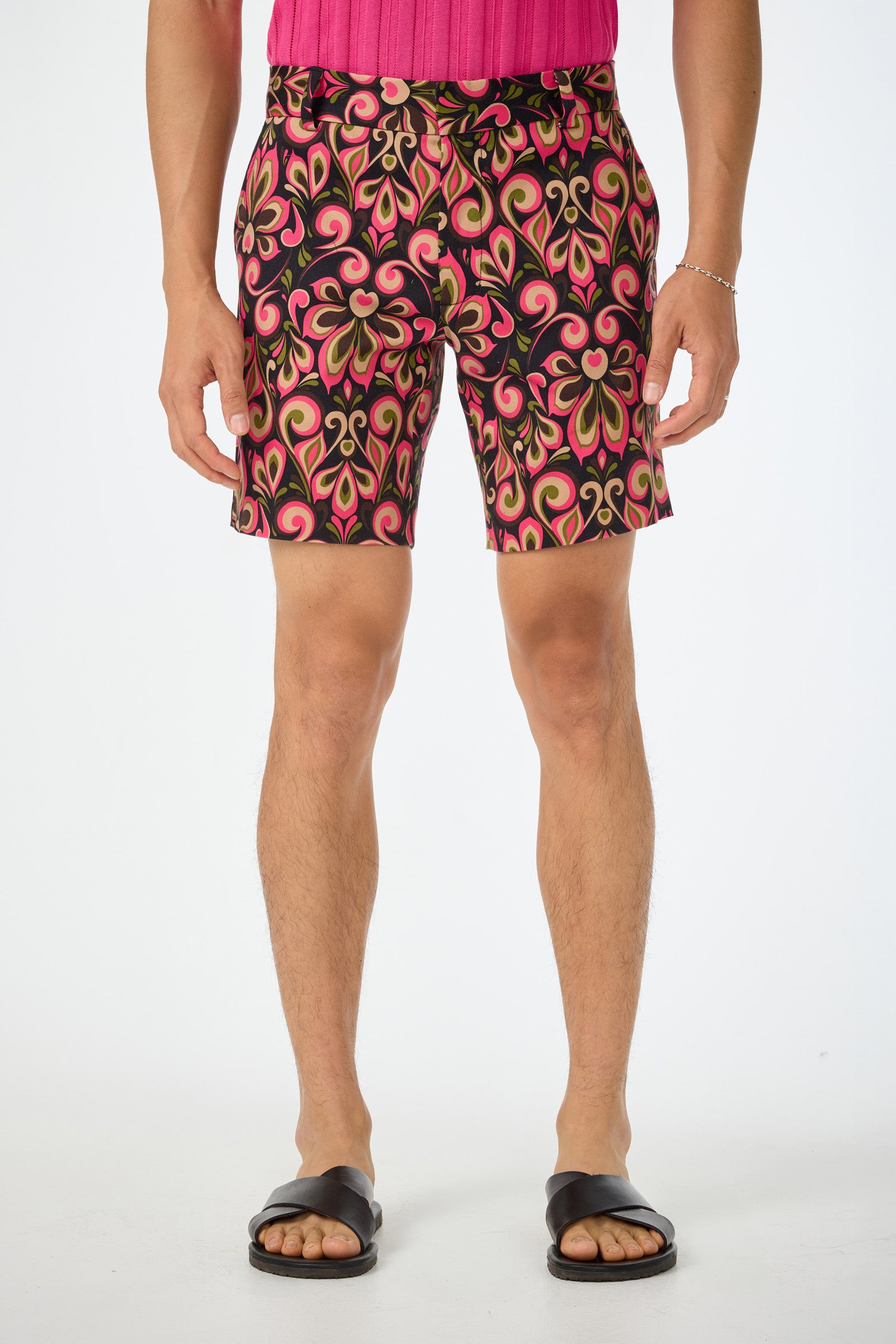 A person wears the Lawrence Short in Sanctuary, featuring a bold pink, red, and green abstract pattern, paired with a pink shirt and black sandals, accessorized with a silver bracelet against a plain white background.