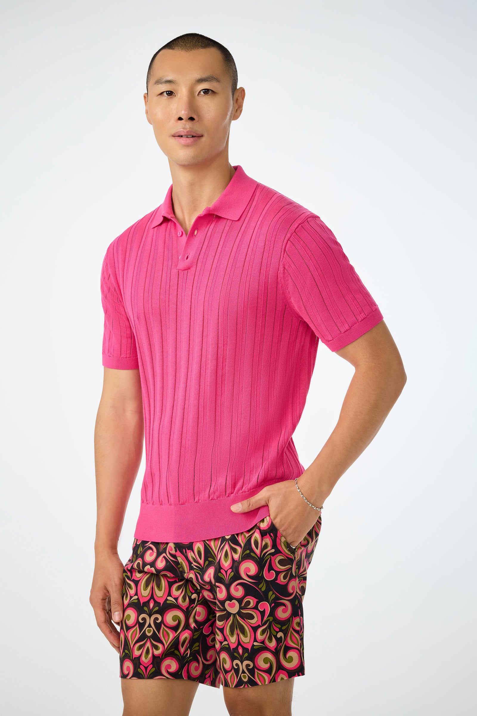 A person stands against a light background wearing the bright pink Kensington Short-Sleeve Polo and colorful patterned shorts with pink, green, and orange swirls, one hand in their pocket.