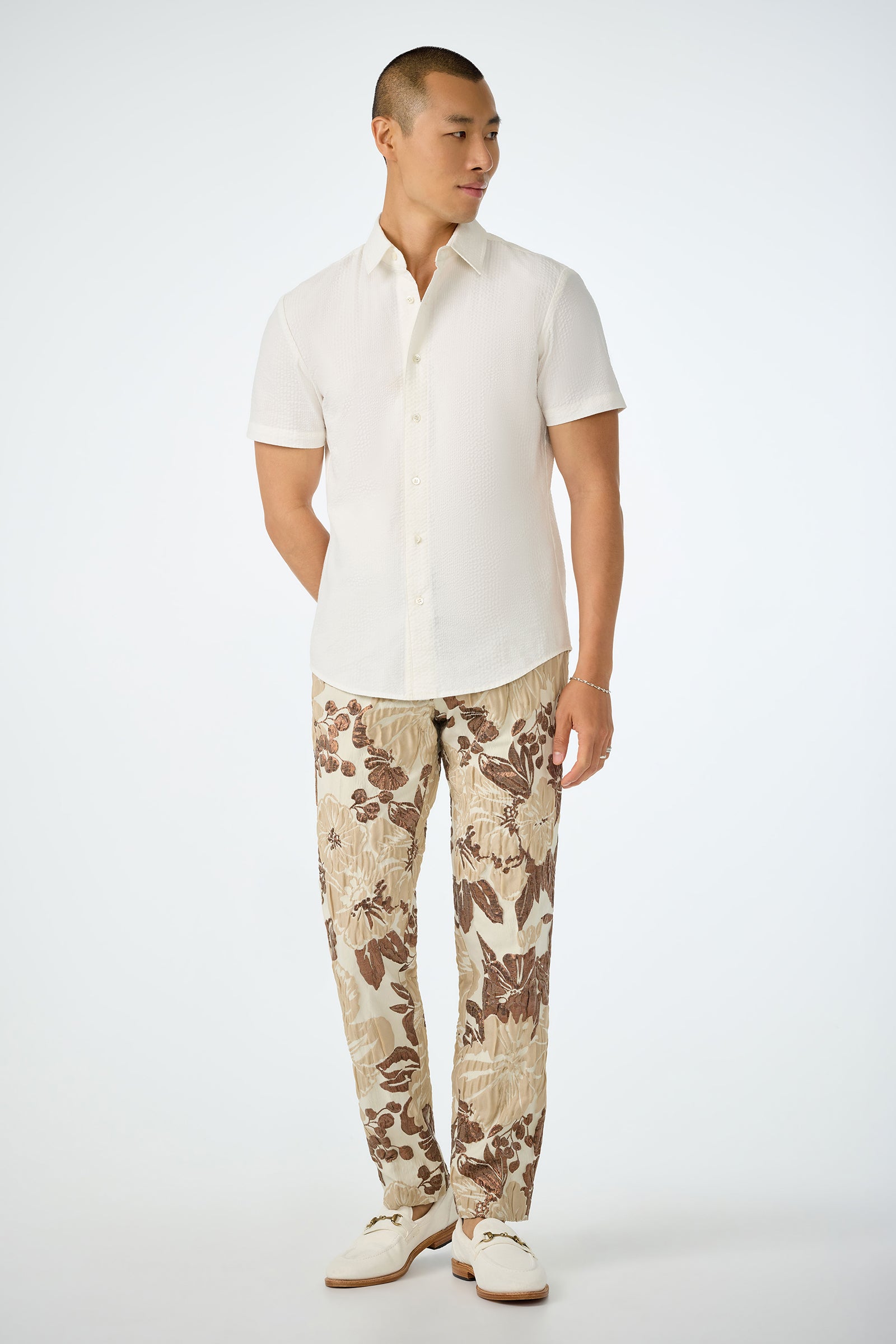 A man stands against a plain background, wearing the Slim Jim Short-Sleeve Shirt in white, beige and brown floral pants, and white shoes. He looks to his left with his hands relaxed by his sides.