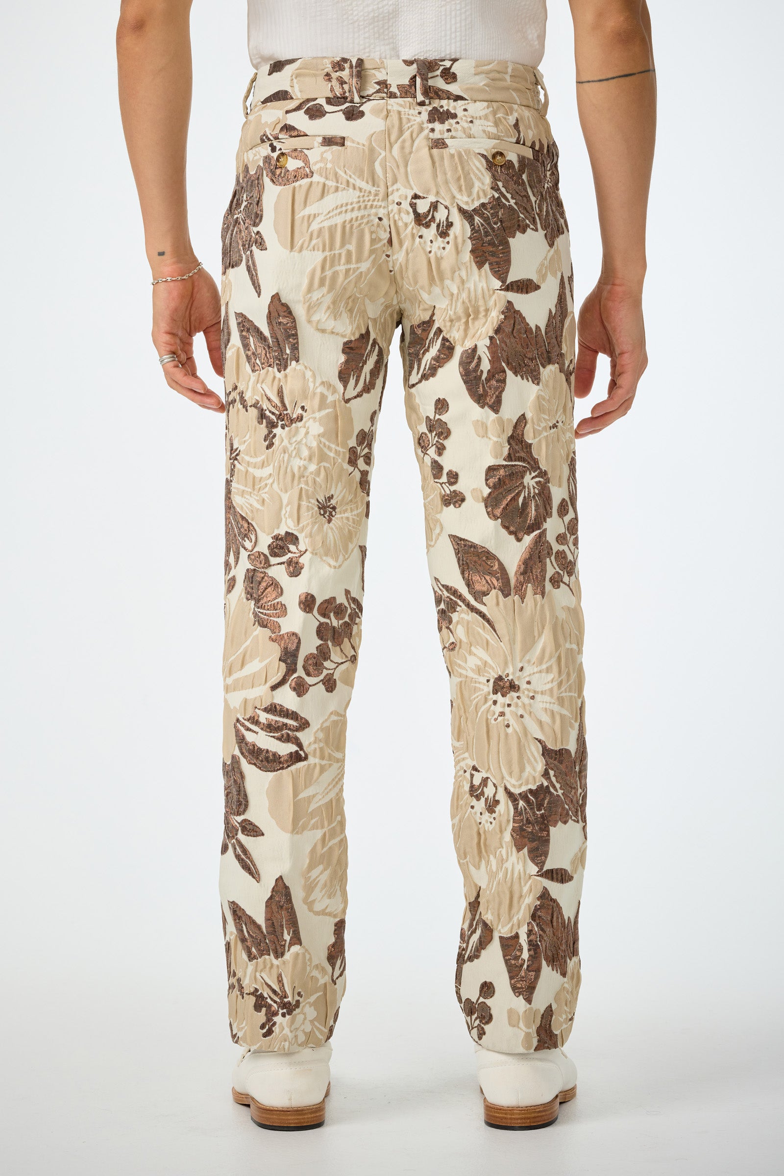 Model stands with back to camera, wearing the Clyde Slim Trouser - Daintree Jacquard featuring a large brown floral pattern, paired with a white top and white shoes. The background is plain white.