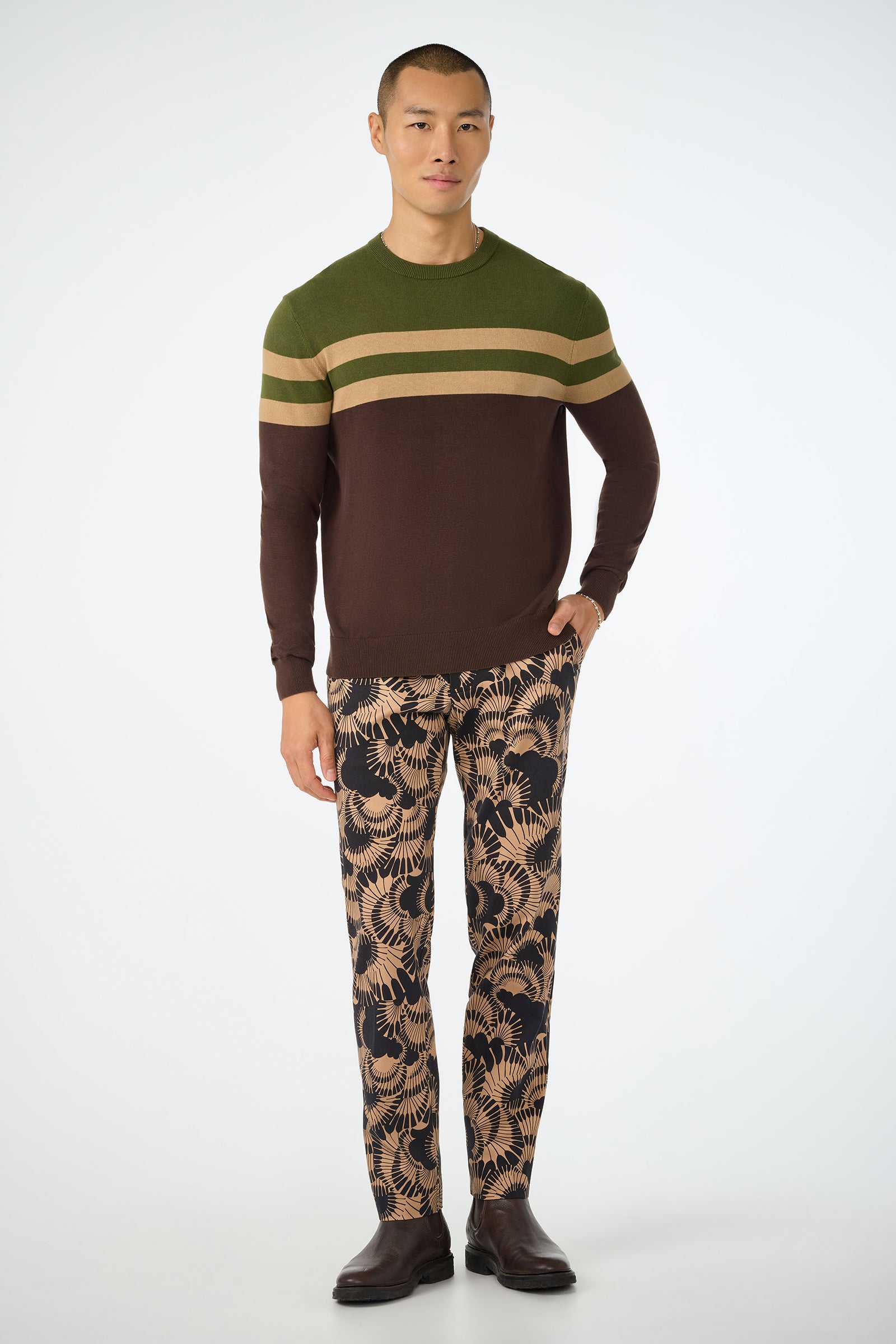 A man stands facing forward in a green and brown Romero Crewneck Sweater with beige stripes, tan and black palm leaf patterned pants, and brown dress shoes against a white background.
