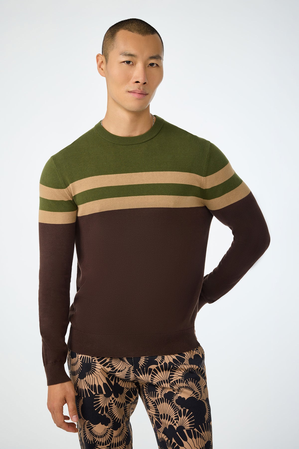 A person stands against a plain background wearing the Romero Crewneck Sweater in multi, featuring olive green, tan, and brown stripes, paired with patterned dark pants with a beige botanical design.
