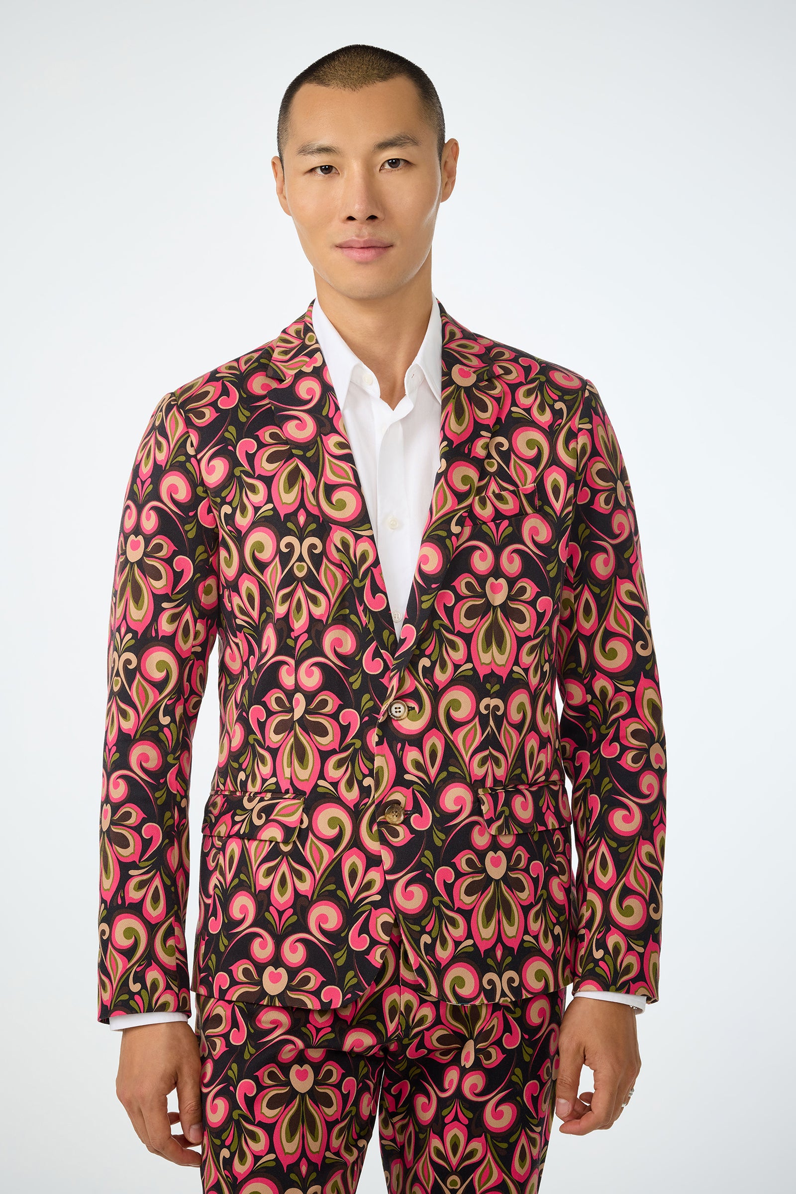 A man with short hair wears the Thurston Blazer in Sanctuary, featuring a bold pink, gold, and black pattern over a white shirt, standing against a plain light background.