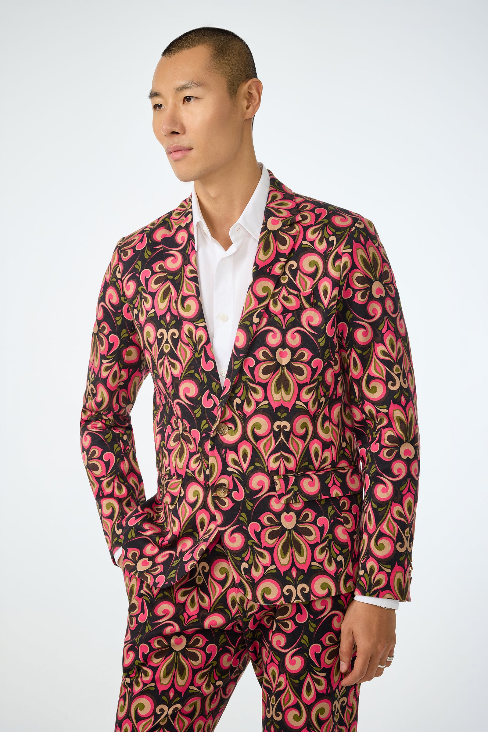 Wearing the Thurston Blazer - Sanctuary, featuring vibrant swirling red, pink, gold, and black patterns over a white shirt, a person stands against a plain white background and gazes to the side with a neutral expression.