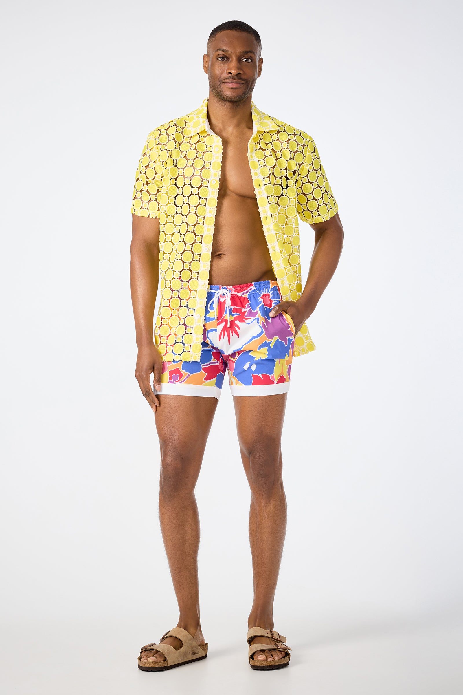 A man stands confidently against a white background, wearing a yellow patterned short-sleeve shirt, tan sandals, and the Hermosa Beach Board Short - Soie Paradis with colorful floral prints.