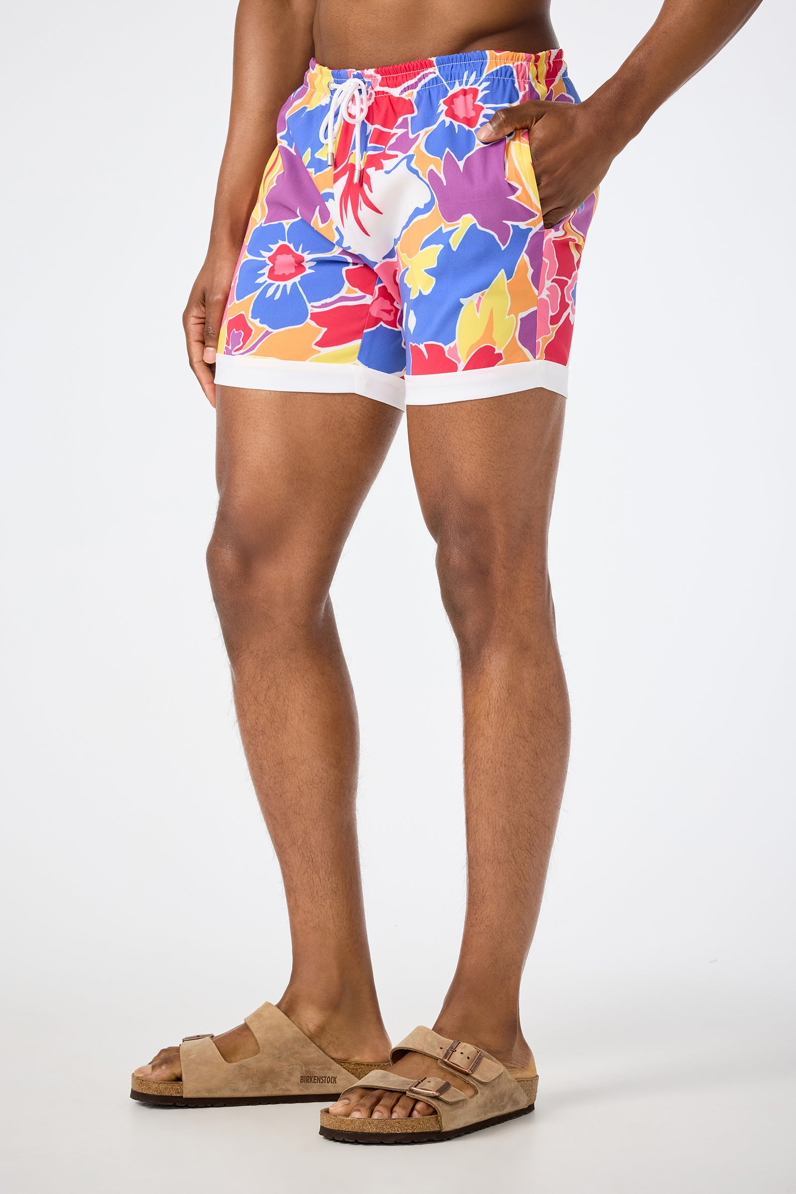 A person models the Hermosa Beach Board Short - Soie Paradis, featuring vibrant floral prints in red, blue, yellow, and purple with white trim, paired with tan Birkenstock-style sandals against a plain white background.