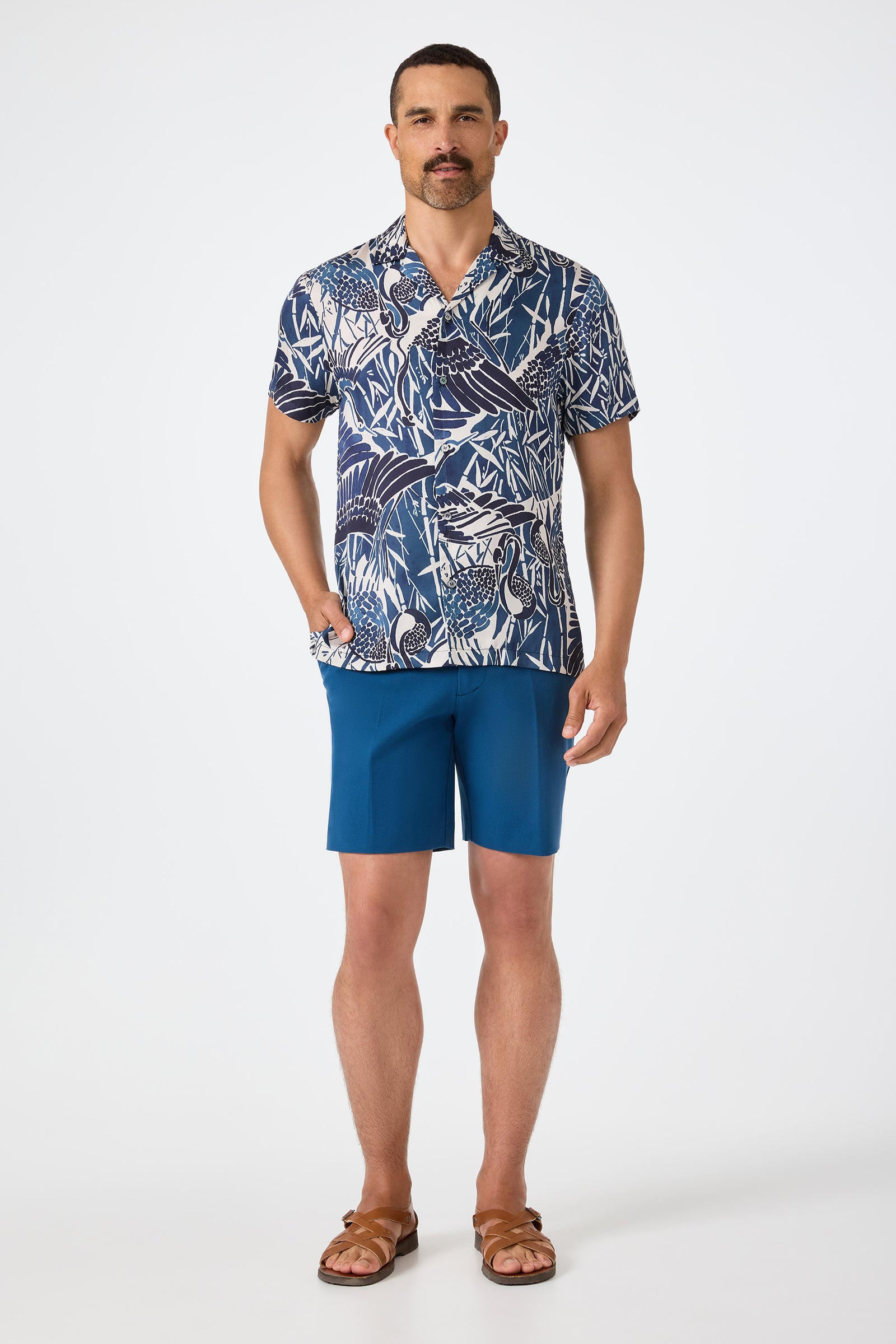 A man with short hair and a mustache stands against a plain background, wearing the Felix Camp Shirt - Tsuru (a blue and white patterned short-sleeve shirt), blue shorts, brown sandals, and has one hand in his pocket.