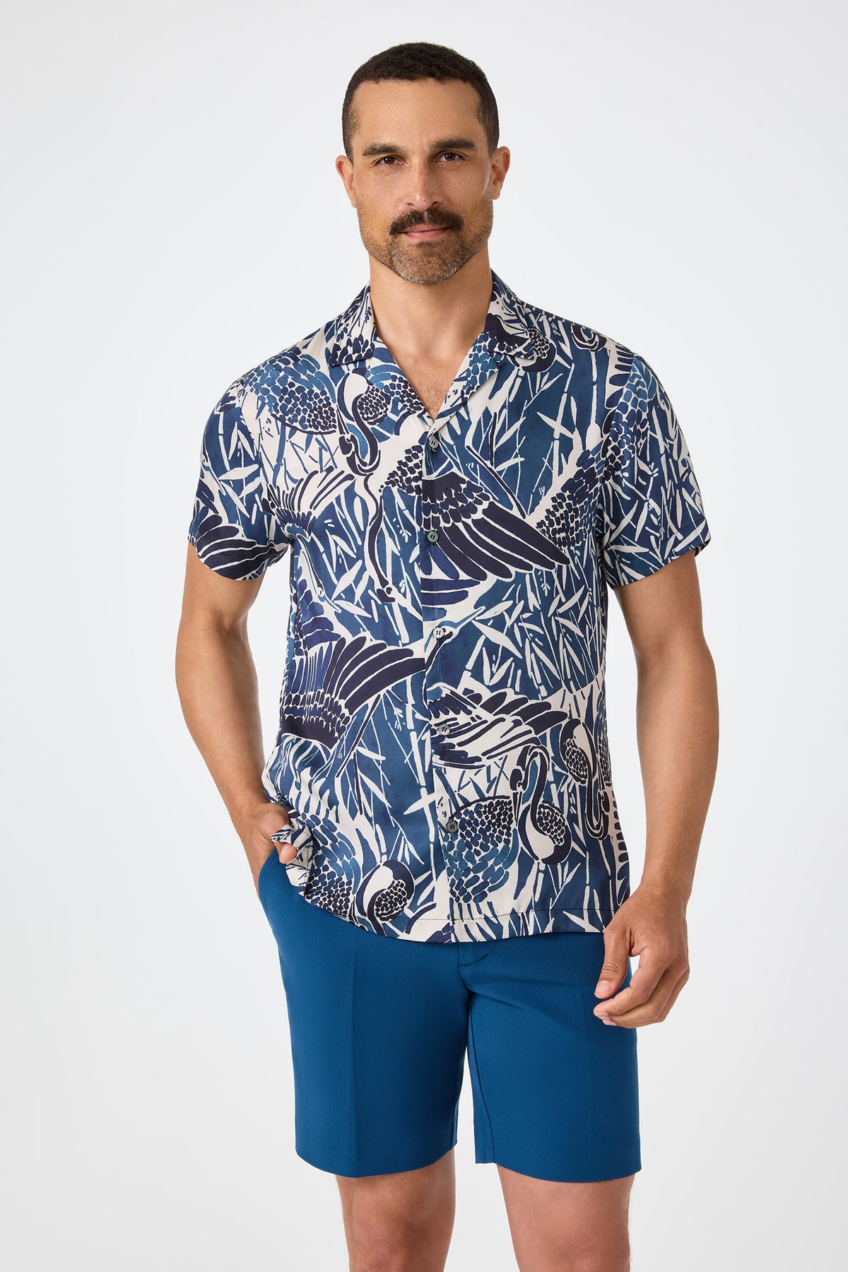 A man with short dark hair and a mustache wears the Felix Camp Shirt in Tsuru, featuring a blue and white pattern, along with solid blue shorts, standing with one hand in his pocket against a plain white background.