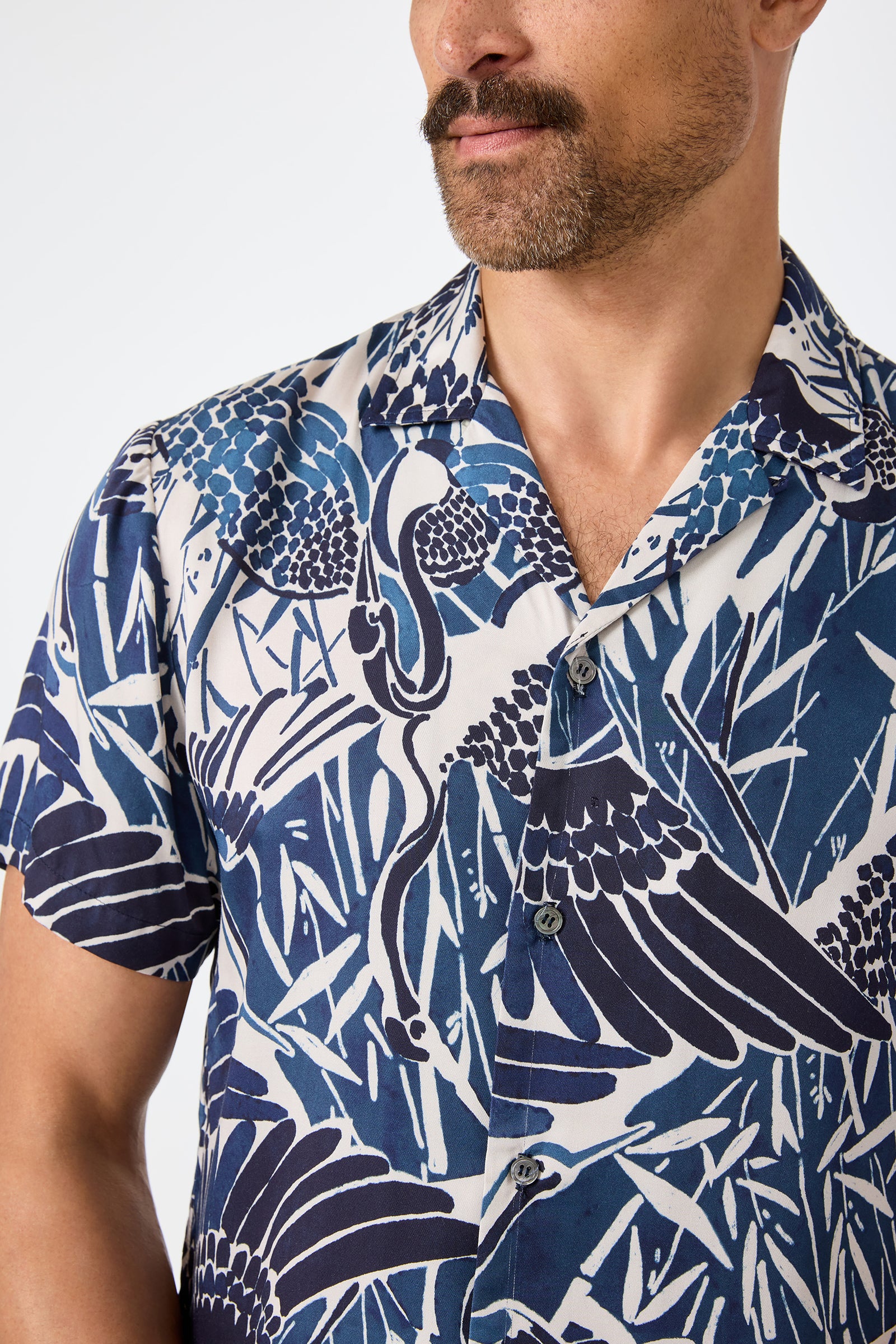 A man with a mustache wears the Felix Camp Shirt - Tsuru, a blue and white short-sleeve shirt with a bold abstract bird and plant pattern. The image is cropped at the shoulders, obscuring his full face.