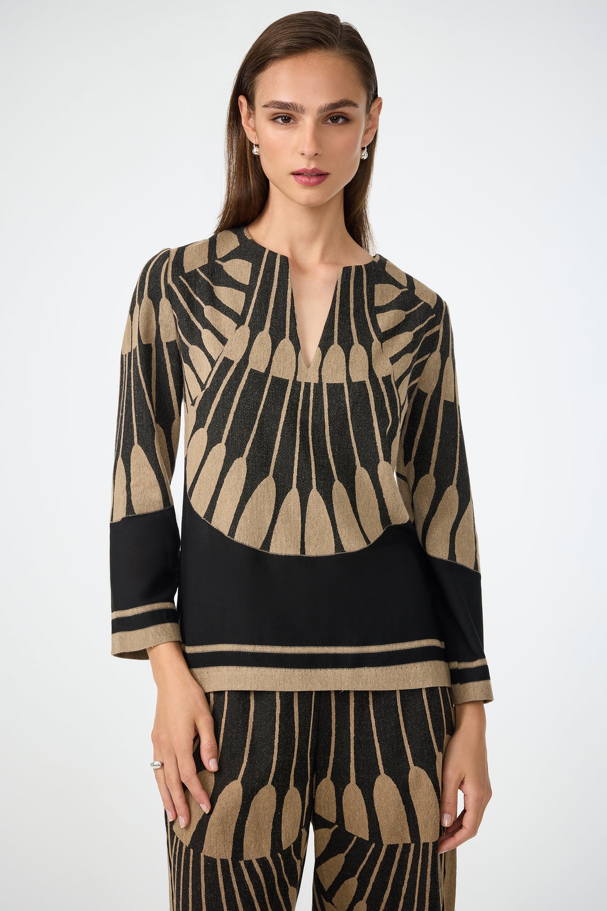 A woman with long brown hair wears the Amy 3/4 Sleeve Top in Juniper Fil Coupe, featuring a black and beige geometric pattern with a V-neck and 3/4 sleeves. She is standing against a plain background.