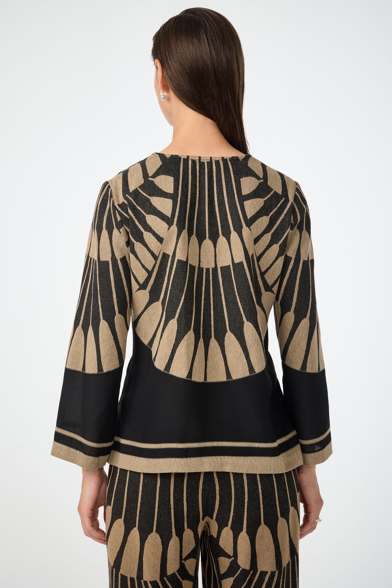 A woman with straight, loose brown hair stands facing away, wearing the Amy 3/4 Sleeve Top in Juniper Fil Coupe—a coordinated outfit with bold black and beige geometric patterns resembling feathers or scales.