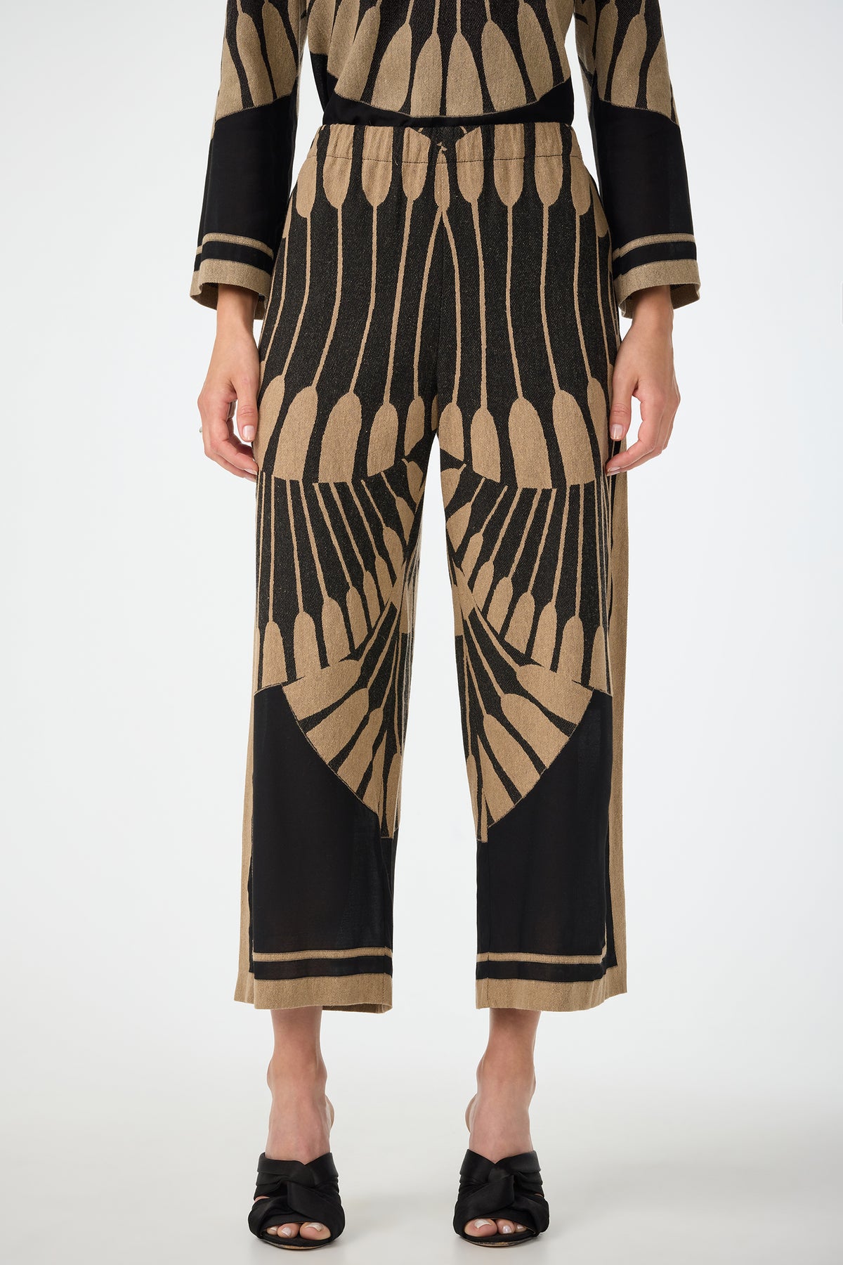 A person wearing Estella 2 Cropped Pants in Juniper Fil Coupe, featuring a bold abstract pattern, and black sandals. Image is cropped from shoulders down on a plain background.