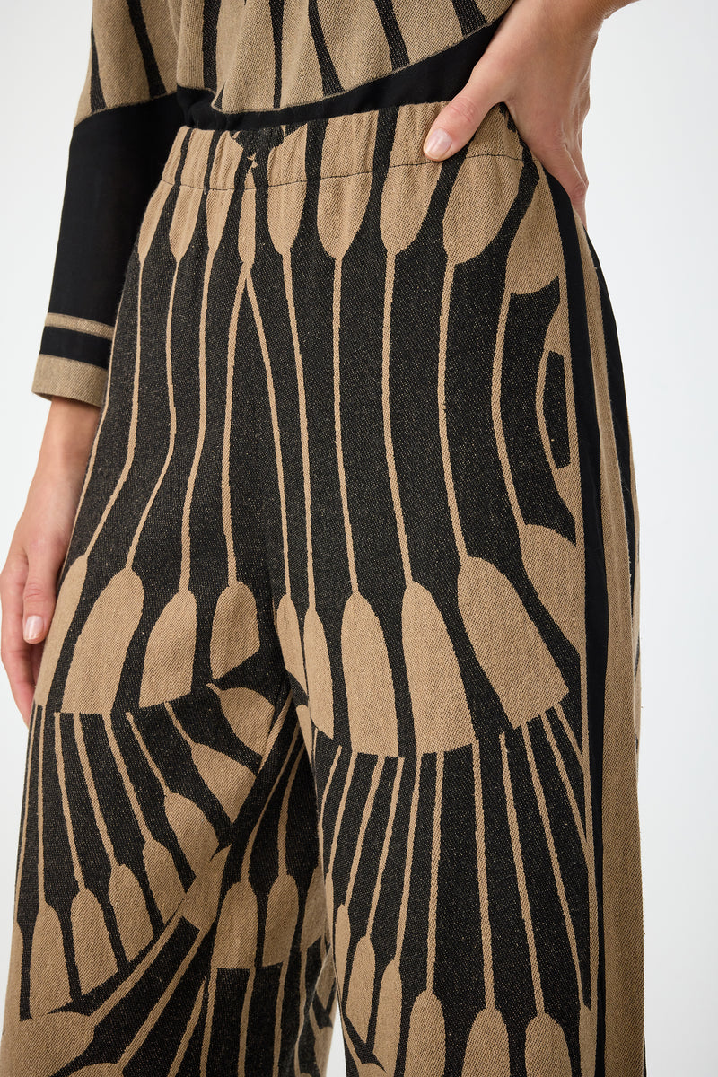 A close-up of a person wearing the Estella 2 Cropped Pants in Juniper Fil Coupe, featuring a bold black and tan abstract pattern. Their hand rests on their hip, and part of a matching top is visible.