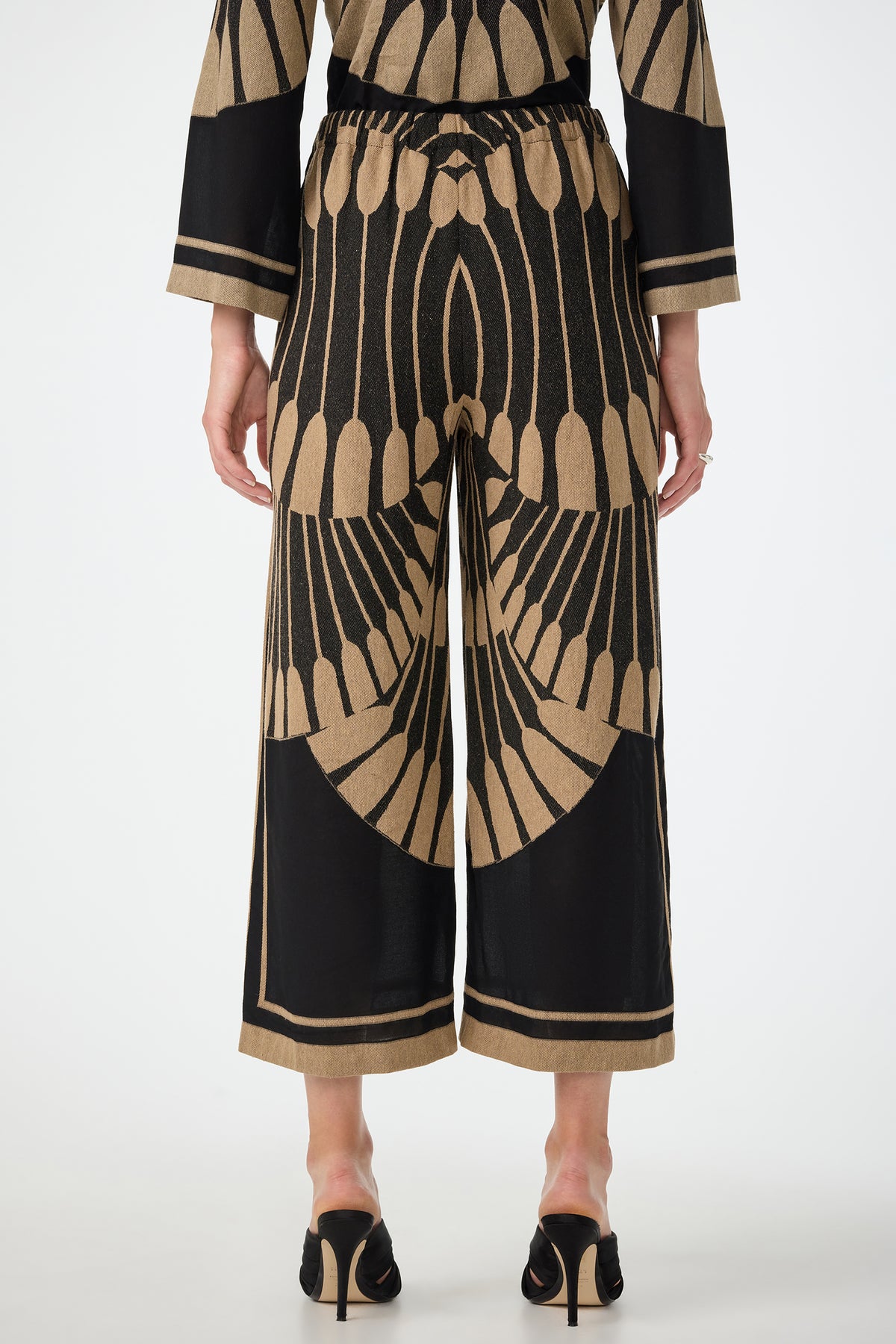 A person is seen from the back wearing Estella 2 Cropped Pants in Juniper Fil Coupe, featuring a wide-leg silhouette and bold tan geometric pattern, paired with a matching top and black heeled sandals against a plain white background.