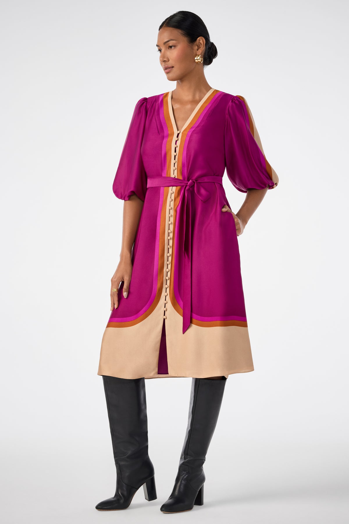 A woman wears the Linden Dress—knee-length, magenta and beige with puffed sleeves, a V-neckline, button details, and a tie belt—paired with black knee-high boots and gold earrings.