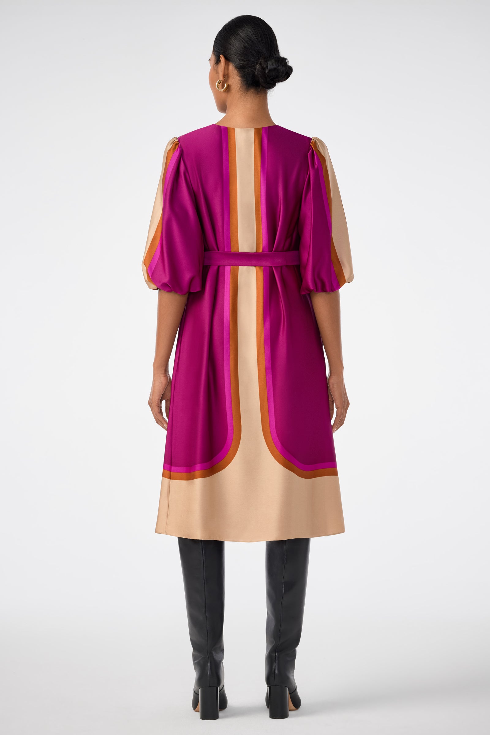 A woman stands facing away, wearing the knee-length Linden Dress with pink, gold, and orange geometric patterns, puffed sleeves, a matching belt, black knee-high boots, and her hair in a low bun.