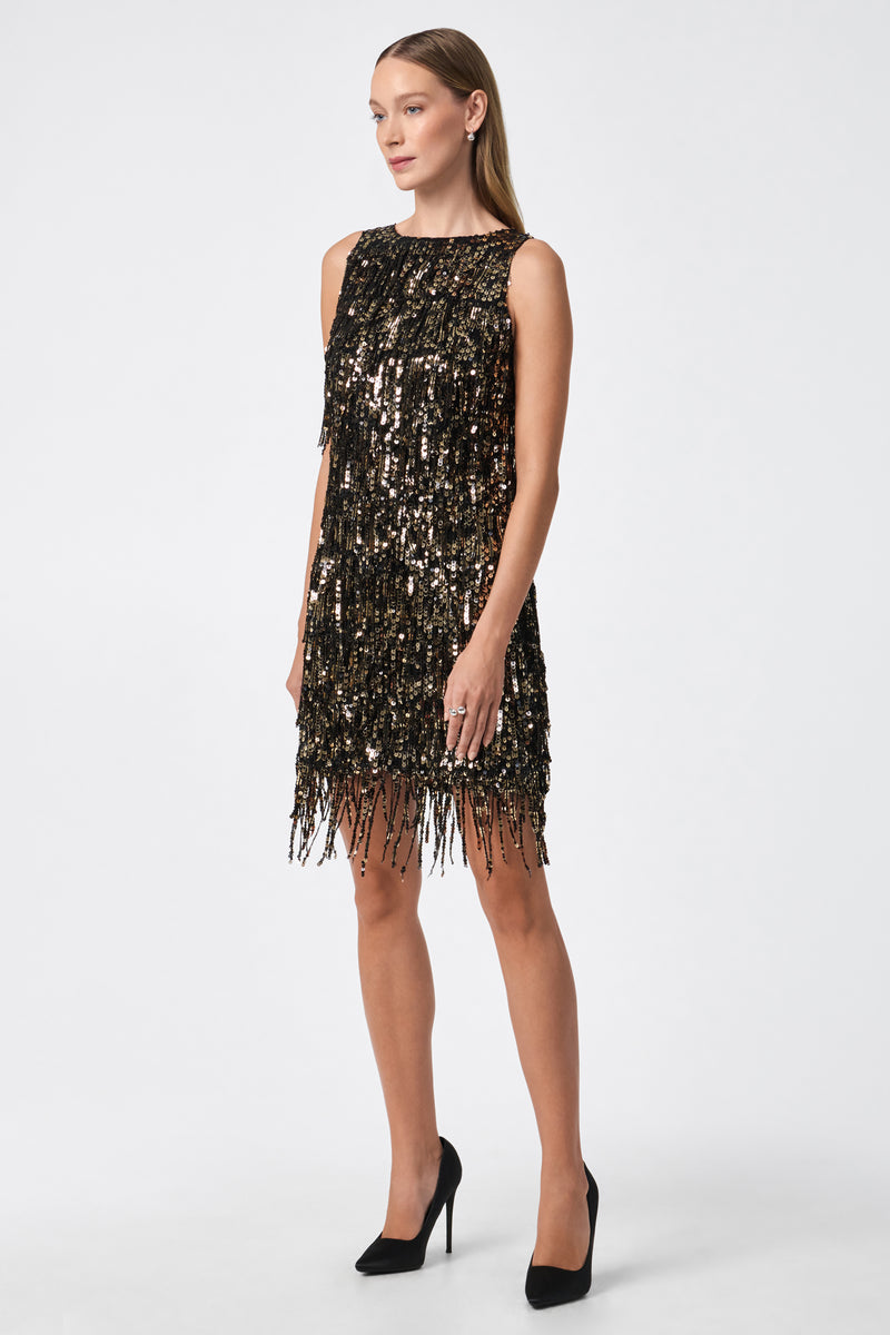 A woman poses against a plain white background wearing the Nini Dress—a sleeveless, knee-length black dress with gold and black sequins, fringe detailing, and paired with black high heels.