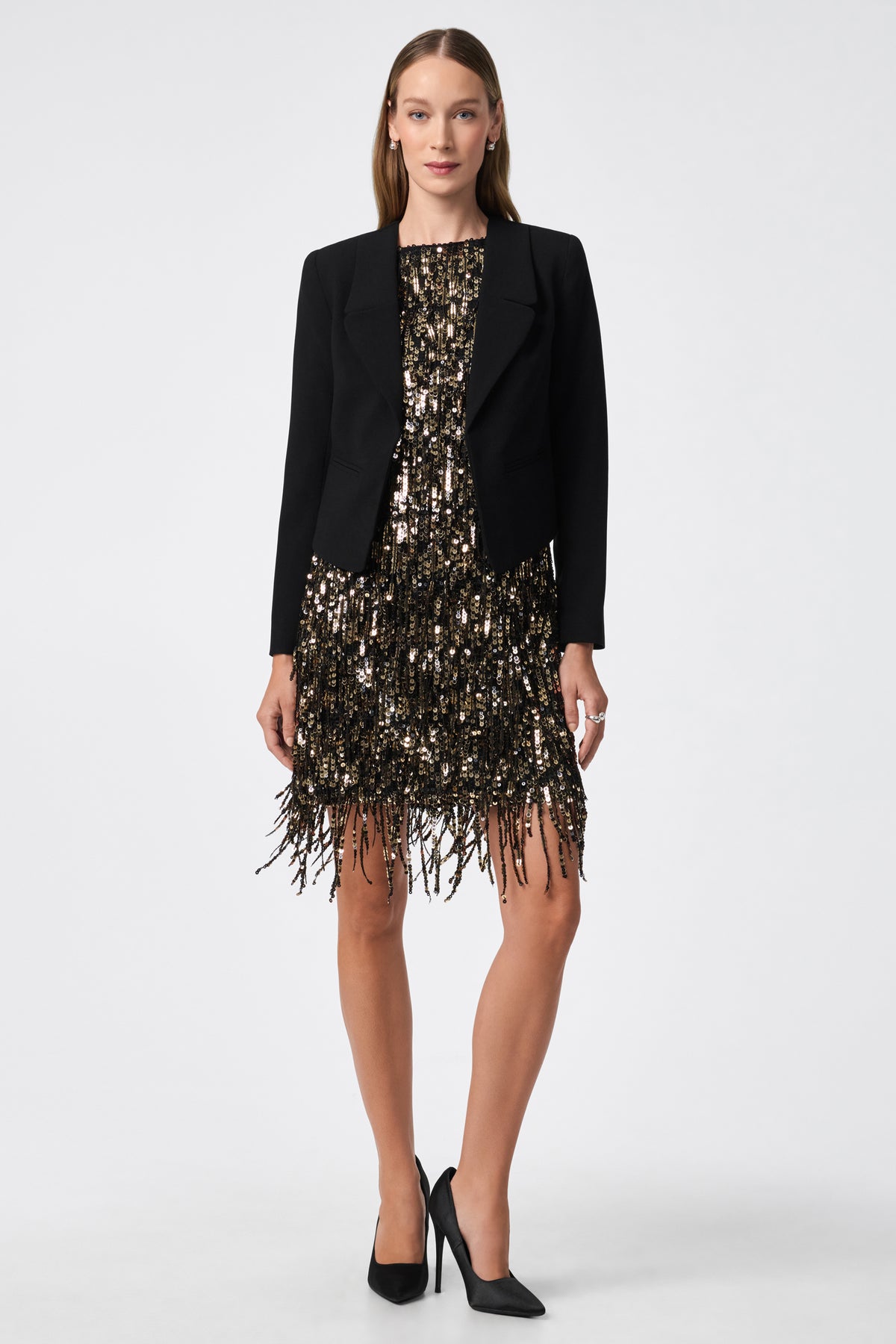 A woman faces forward in a black Ren Cropped Blazer over a short gold and black sequin fringe dress. She has straight brown hair, wears black high heels, and stands against a plain white background.
