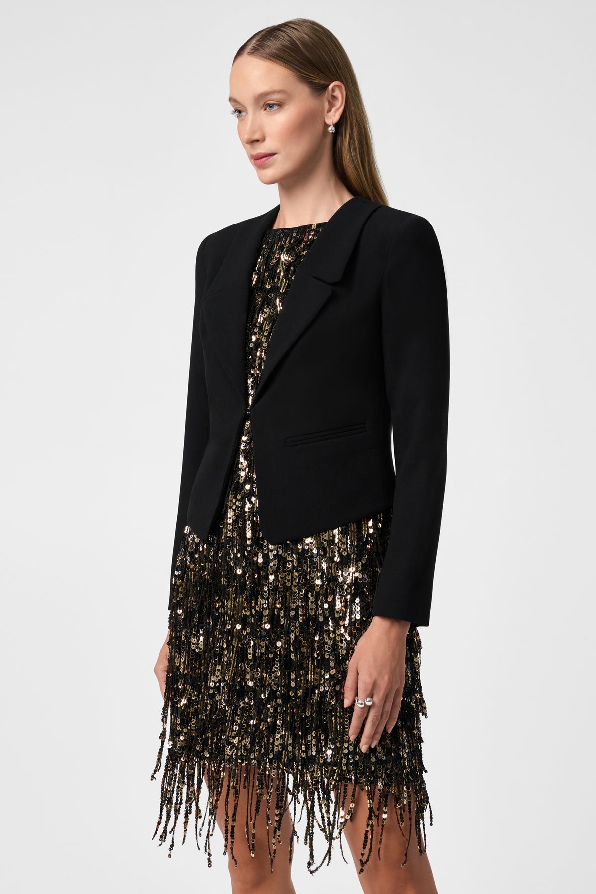 A woman with straight light brown hair poses against a plain white background, wearing the Ren Cropped Long Sleeve Blazer Jacket over a black and gold sequined fringe dress.