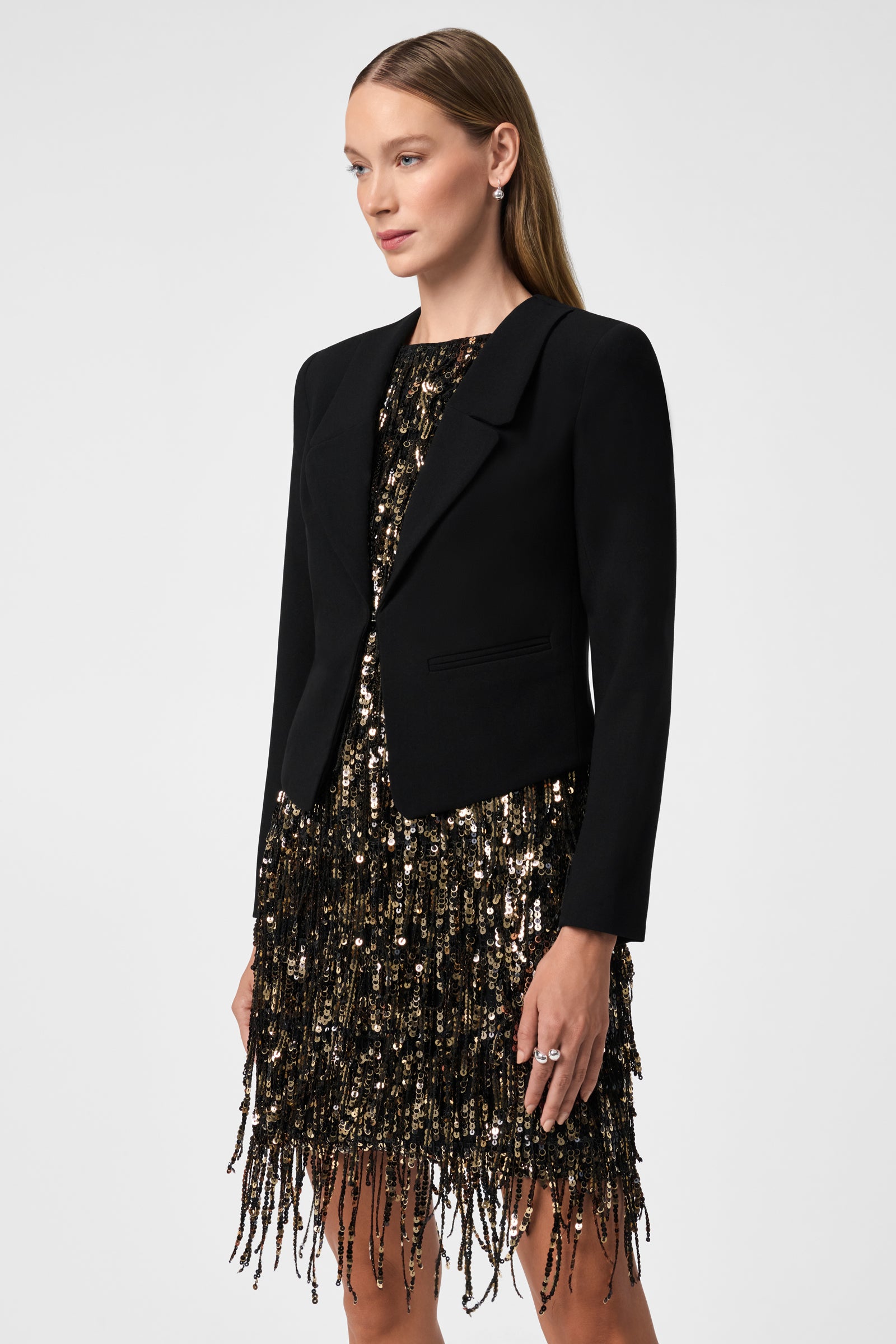A woman with straight light brown hair poses against a plain white background, wearing the Ren Cropped Long Sleeve Blazer Jacket over a black and gold sequined fringe dress.