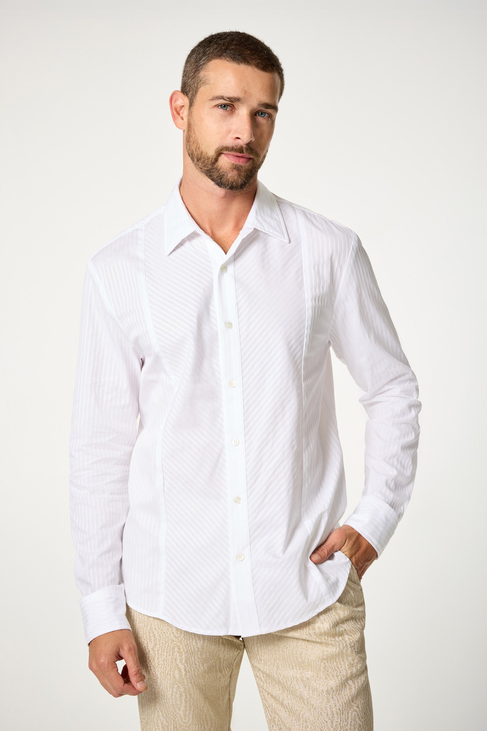 A man with short brown hair and a beard wears the Baleri Tux Shirt, white and buttoned up, paired with beige pants. He stands against a plain light background with one hand in his pocket, looking at the camera with a neutral expression.