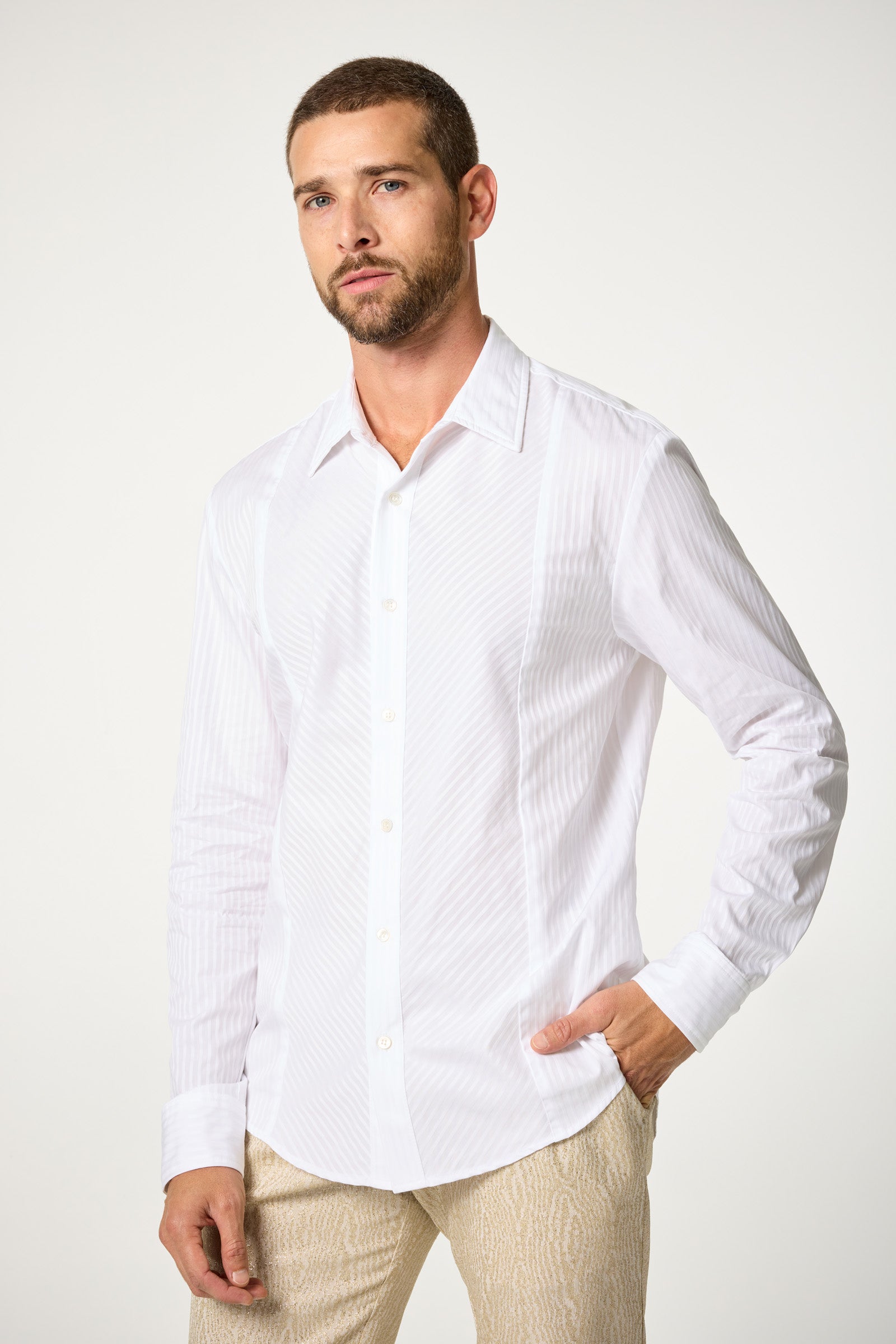 A man with short hair and a beard models the Baleri Tux Shirt, a white, long-sleeved button-up, paired with light pants. He stands against a plain light background, one hand in his pocket, gazing slightly to the side.
