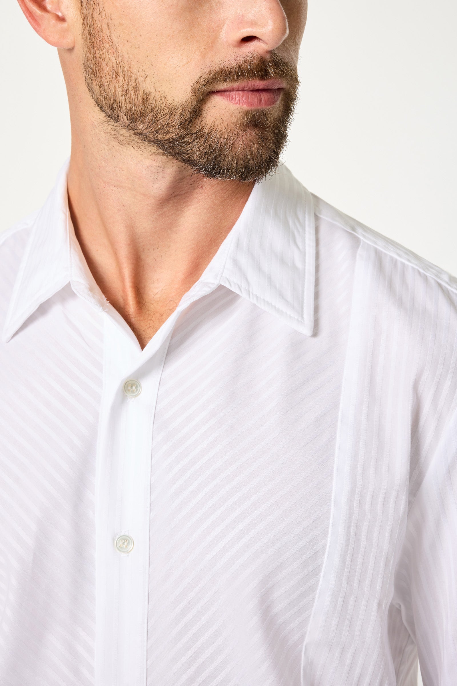 A man with a trimmed beard wears the Baleri Tux Shirt—a white, textured button-up with a pointed collar—while looking slightly to the side. Only his mouth and part of his face are visible.