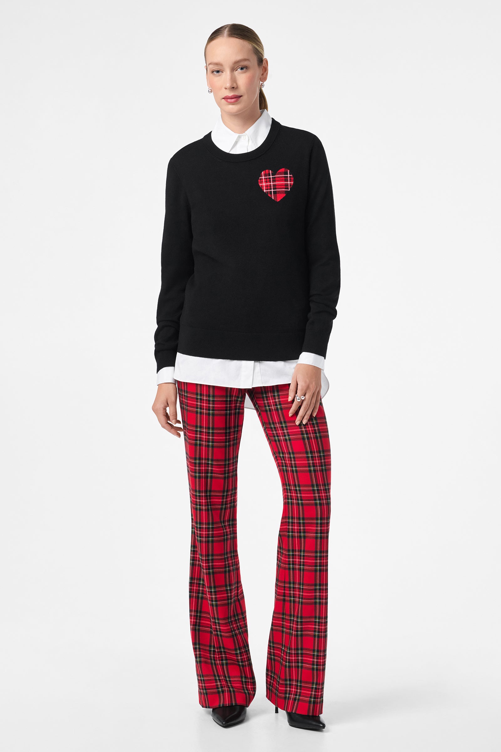 A woman wears the Plaid Heart Sweater, featuring a black knit with a red plaid heart on the chest, layered over a white collared shirt, paired with red plaid flared pants and black shoes against a plain white background.