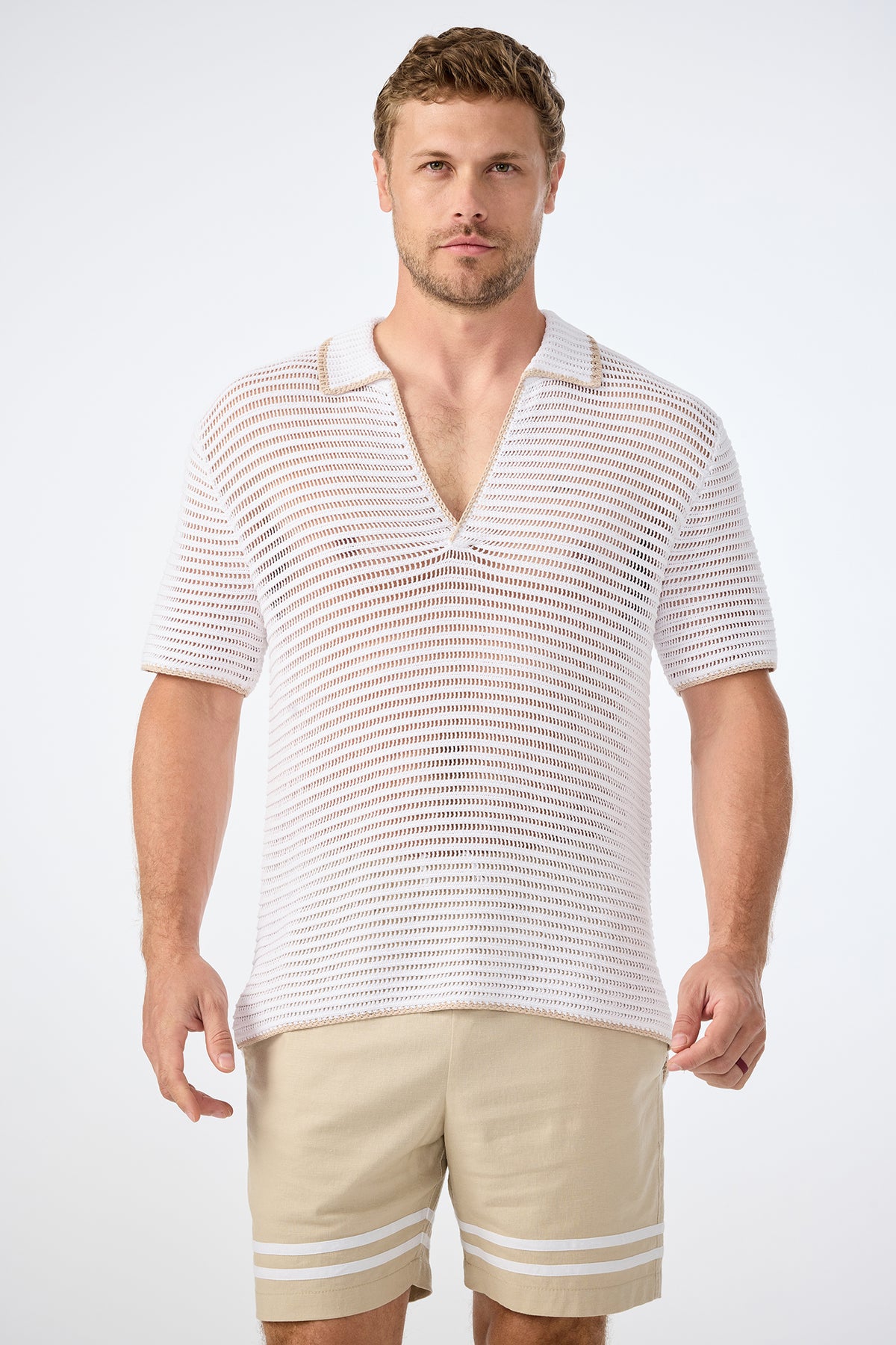 A man faces forward in a plain, light setting, wearing the Triston Short-Sleeve Crochet Polo—white, collared, V-neck and loosely knit—paired with light beige shorts featuring white stripes near the hem.