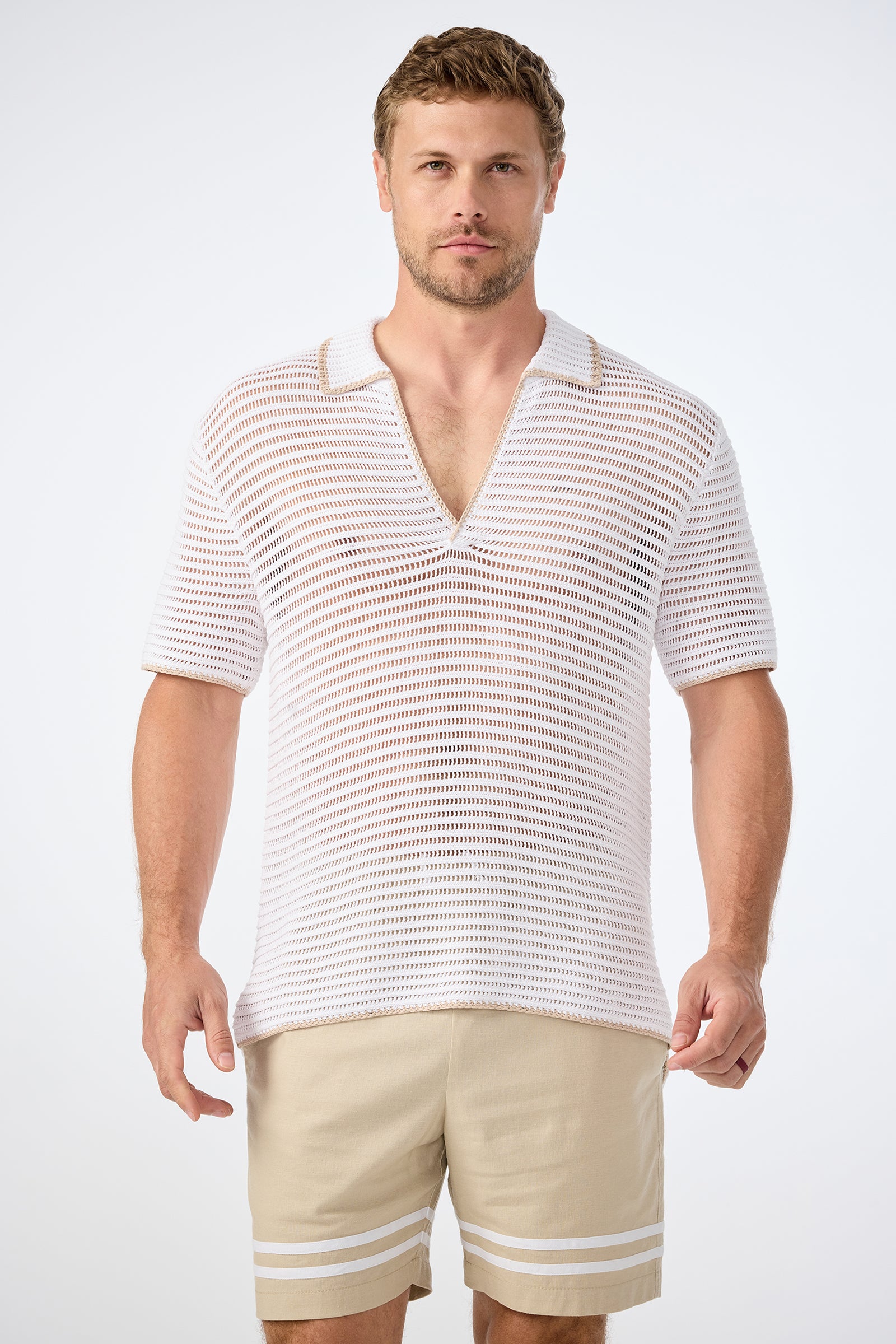 A man faces forward in a plain, light setting, wearing the Triston Short-Sleeve Crochet Polo—white, collared, V-neck and loosely knit—paired with light beige shorts featuring white stripes near the hem.